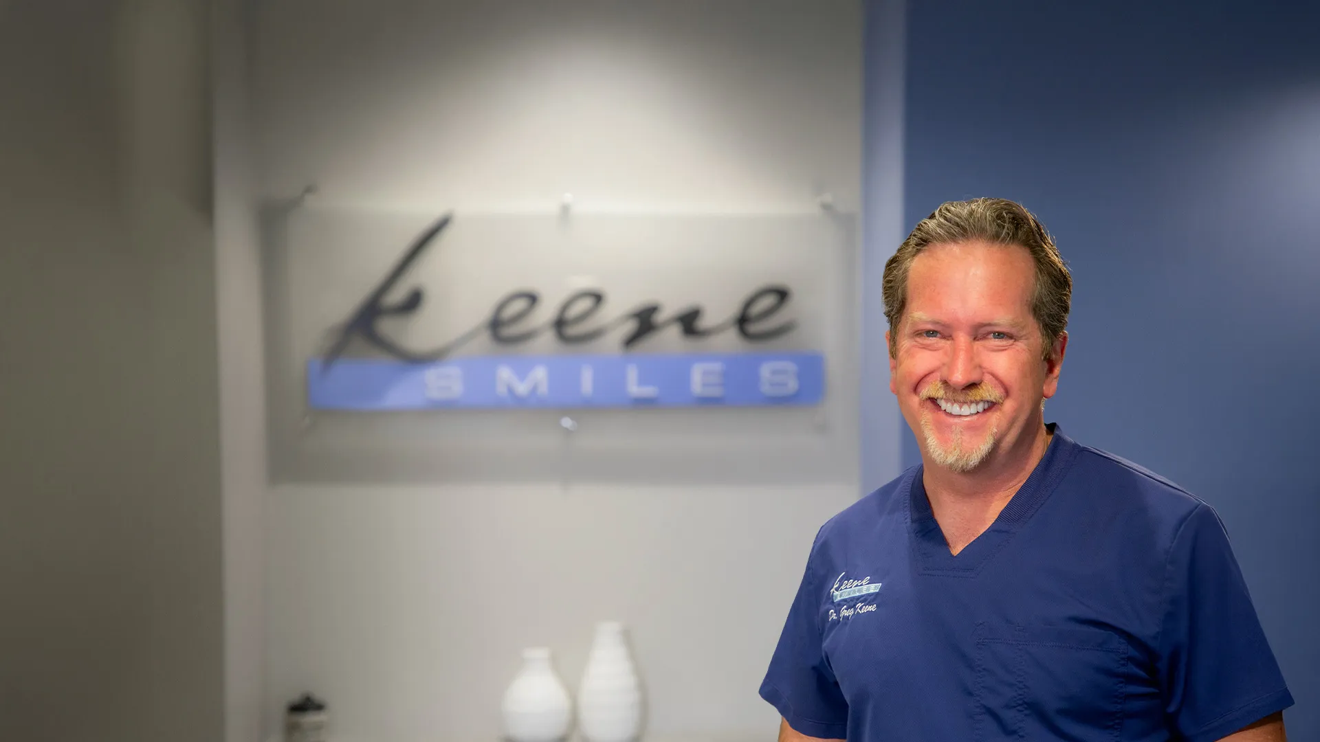 Smiling male dentist in blue scrubs standing in front of Keene Smiles dental office sign.