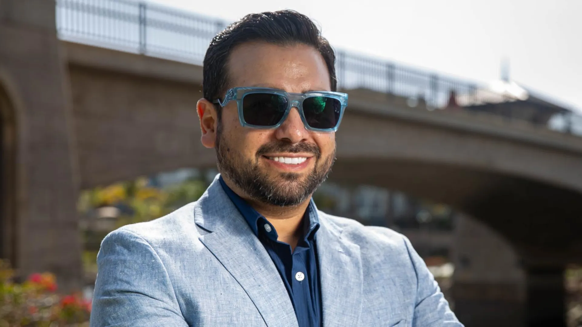 Smiling man in sunglasses poses confidently by a bridge on a sunny day.