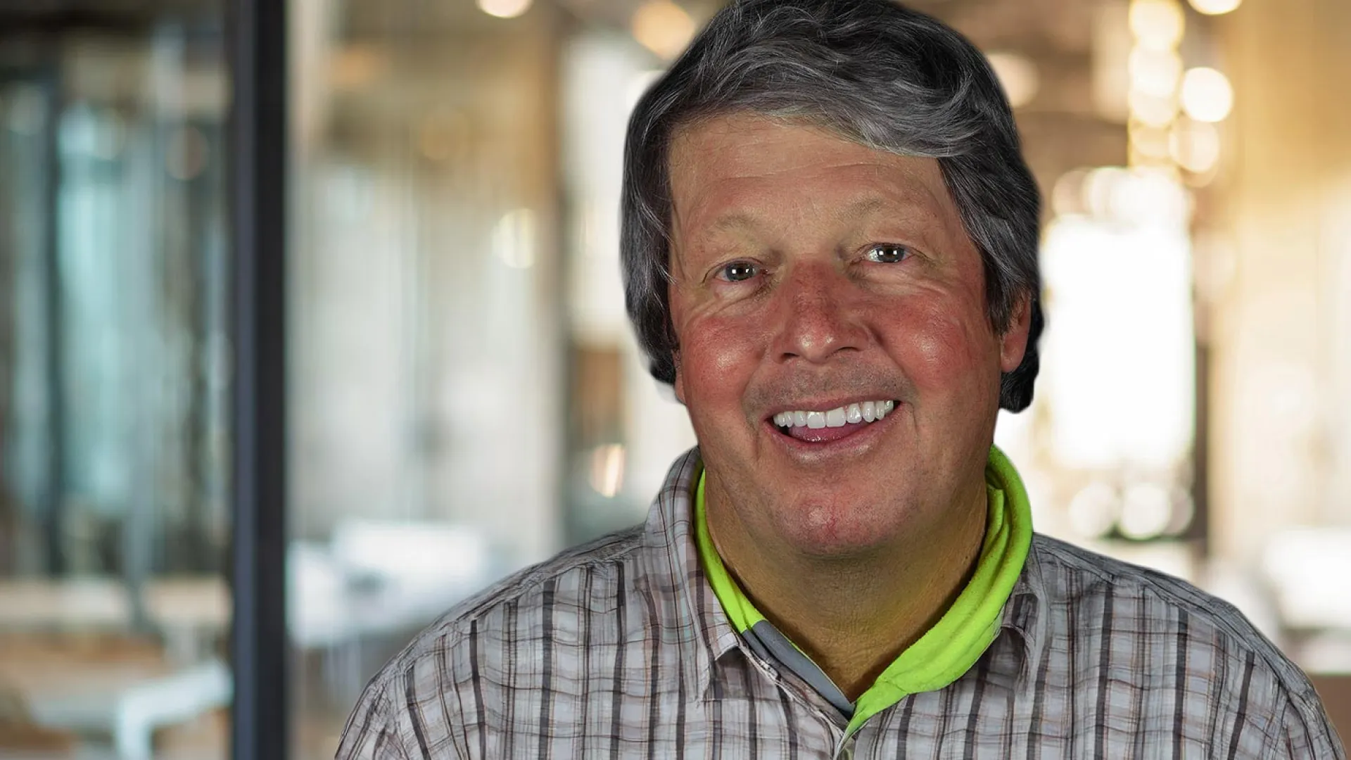 Smiling man in a checked shirt with a modern office backdrop and warm lighting.