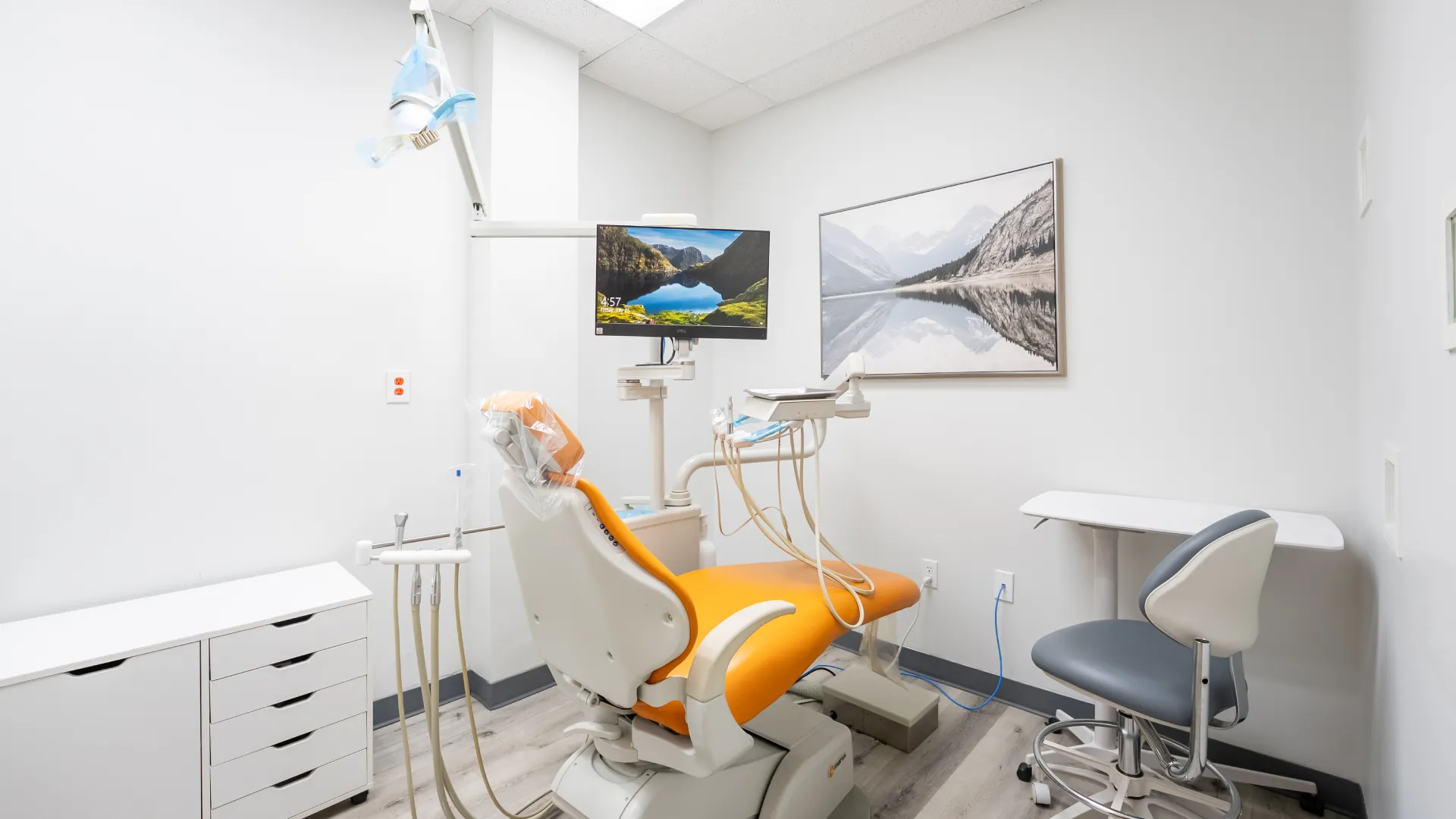 Modern dental office with orange patient chair, dental equipment, monitor, and nature-themed wall art in white room.