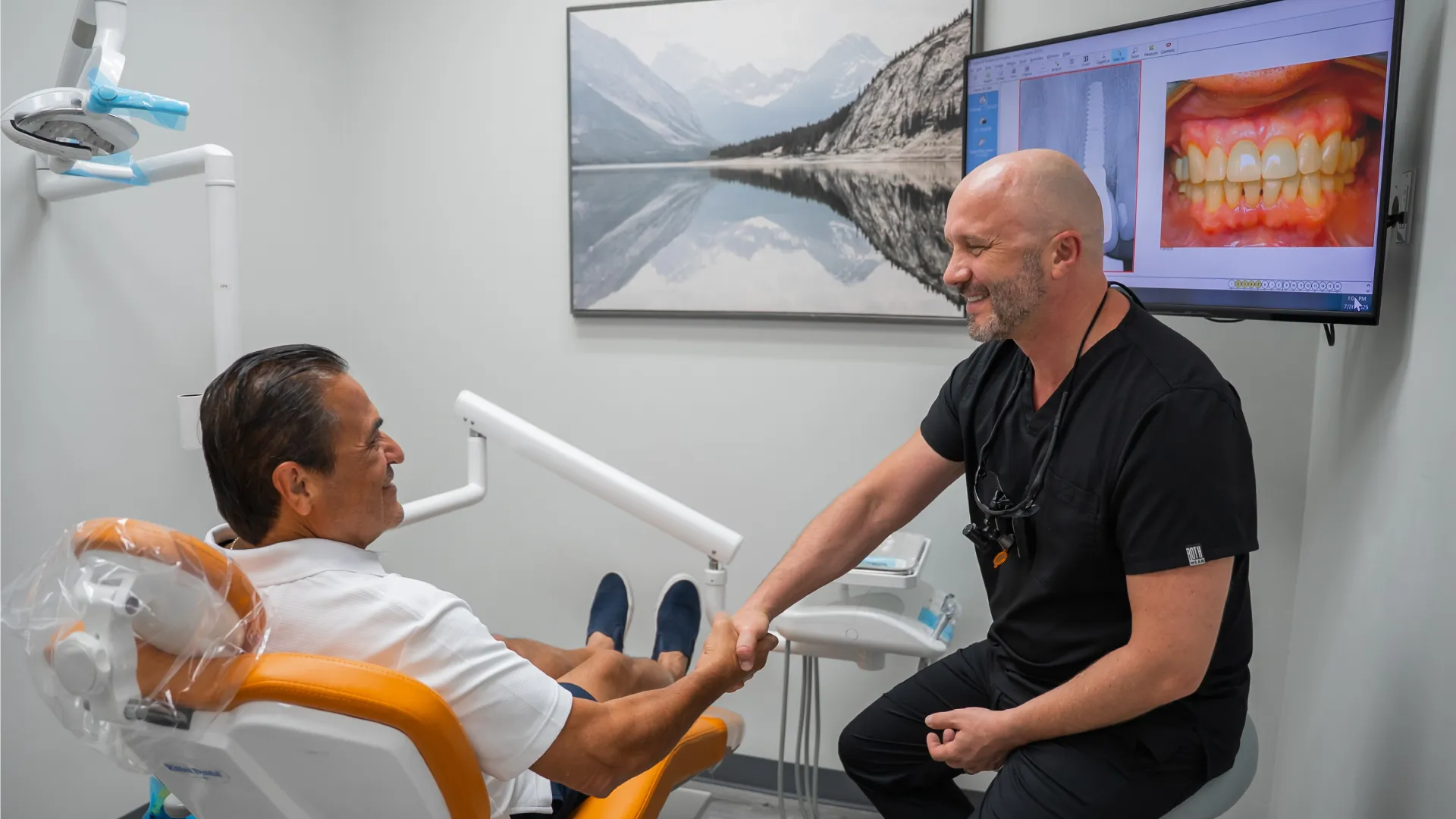 Dentist shaking hands with patient in dental office with teeth images on screen and mountain photo on wall