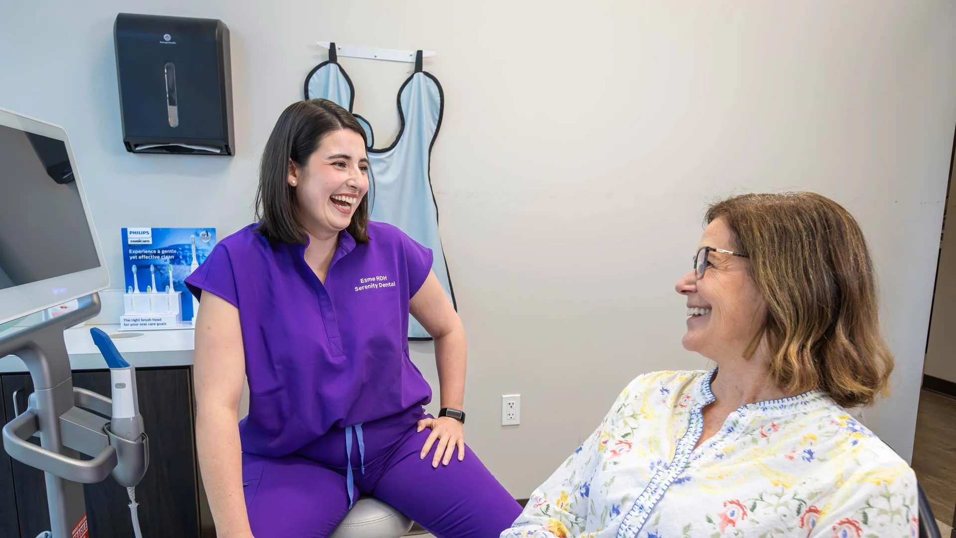Dentist in purple scrubs smiling and talking with a female patient in a dental office consultation room.