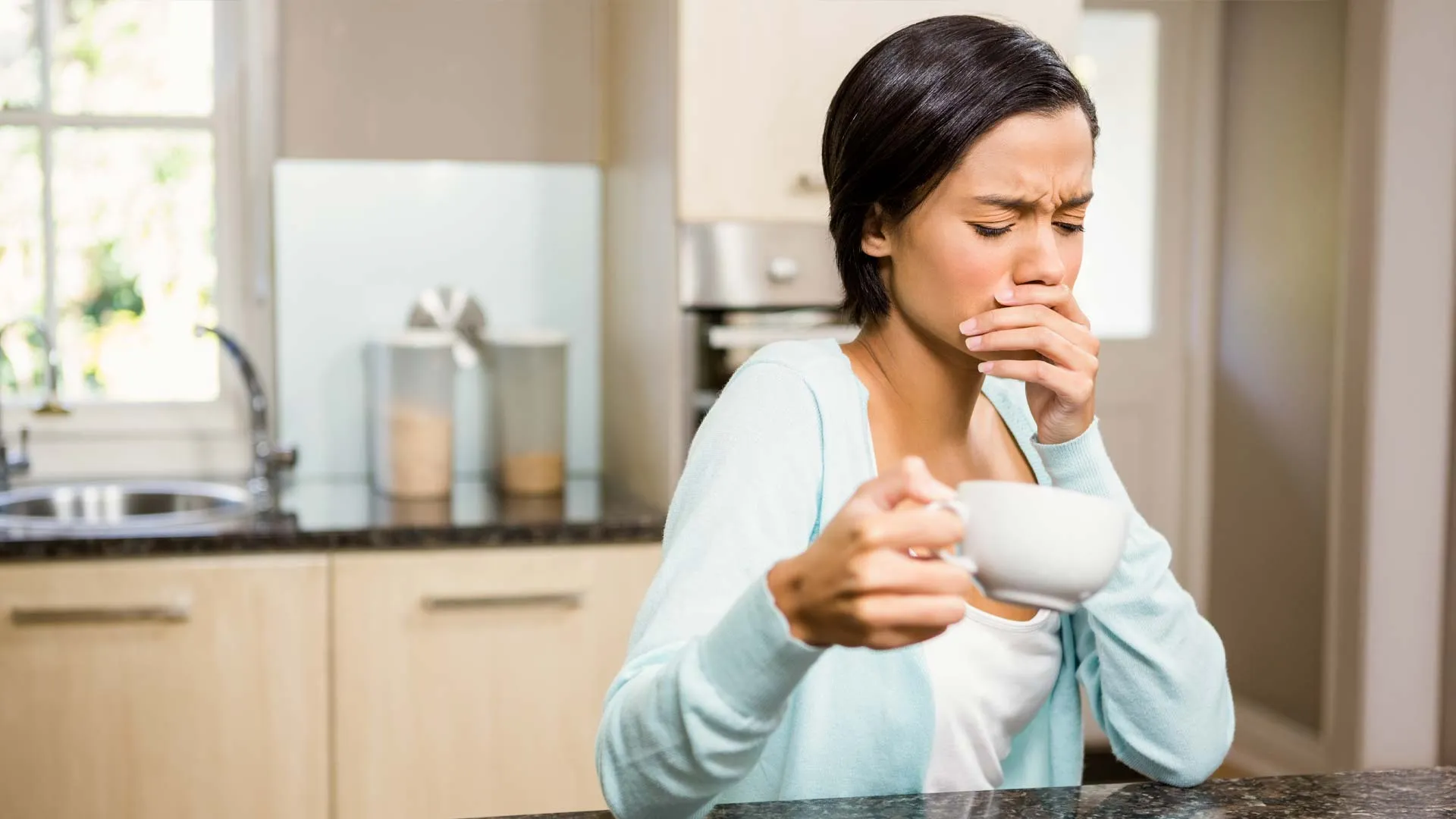 Young woman in kitchen coughing while holding a white cup and covering her mouth with her hand.