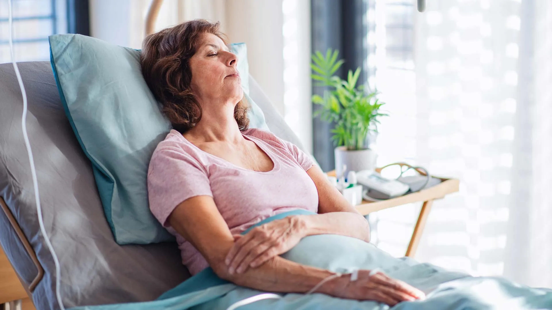 Middle-aged woman resting with eyes closed in a hospital bed with IV drip in a bright room.