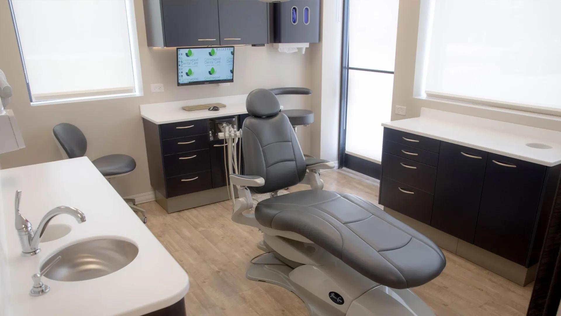 Modern dental office with reclining patient chair, cabinetry, sink, and wall-mounted monitor in bright room.