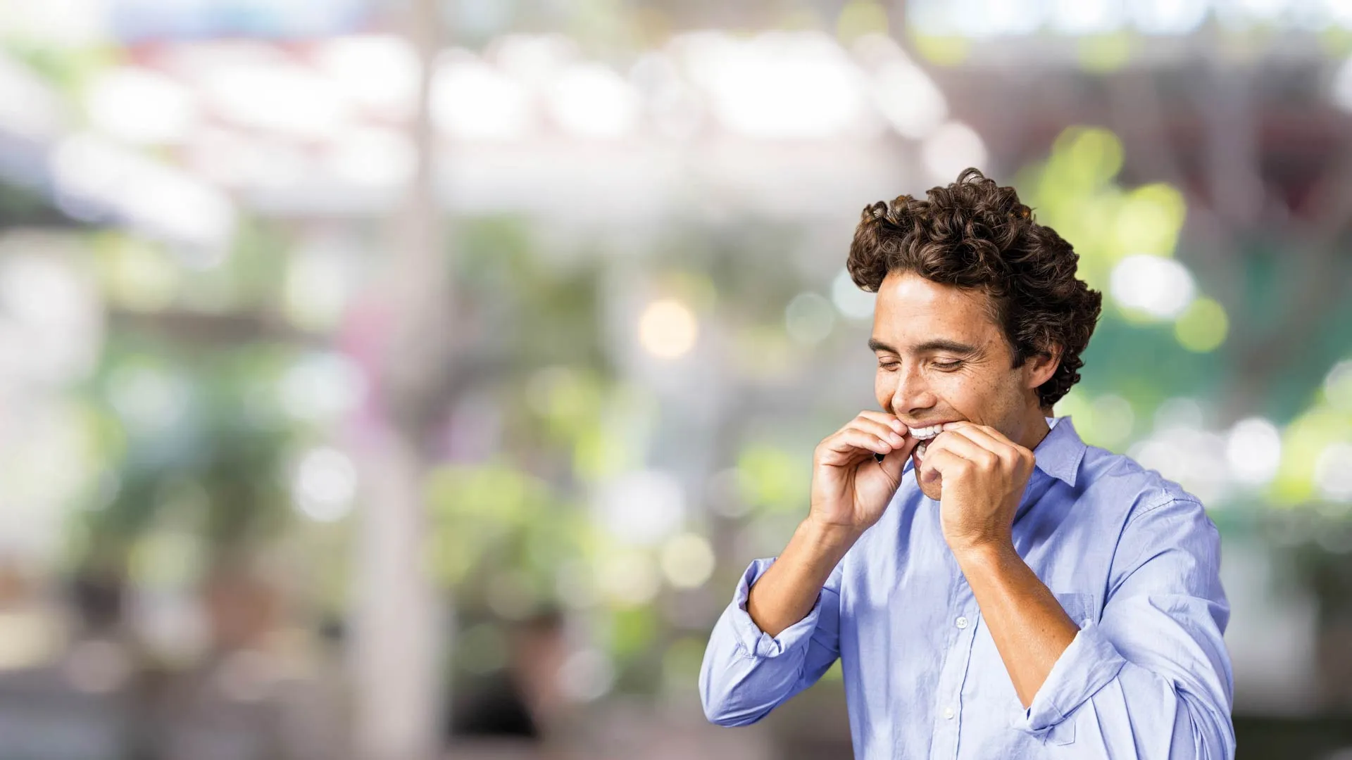 Man in light blue shirt smiling and flossing his teeth with blurred outdoor background.