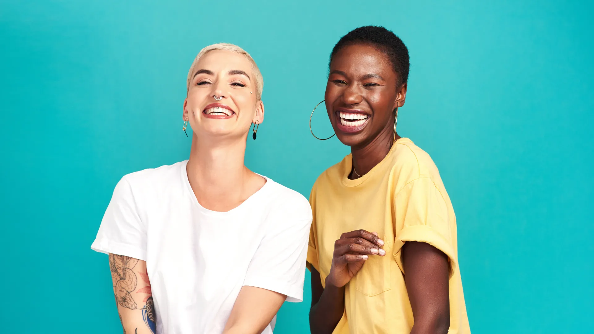 Two young women smiling and laughing against a bright teal background, one with tattoos and short blonde hair, the other with short black hair.
