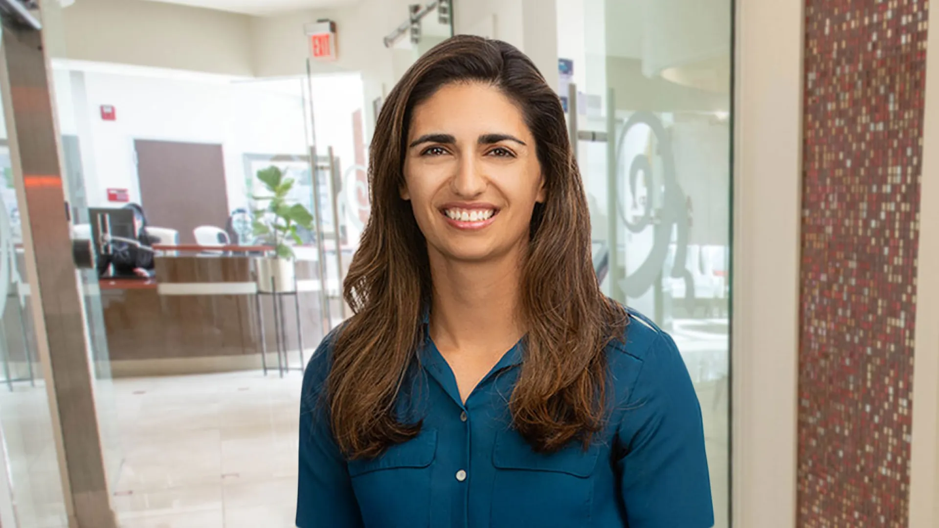 Smiling woman in a teal shirt stands in a modern office lobby.