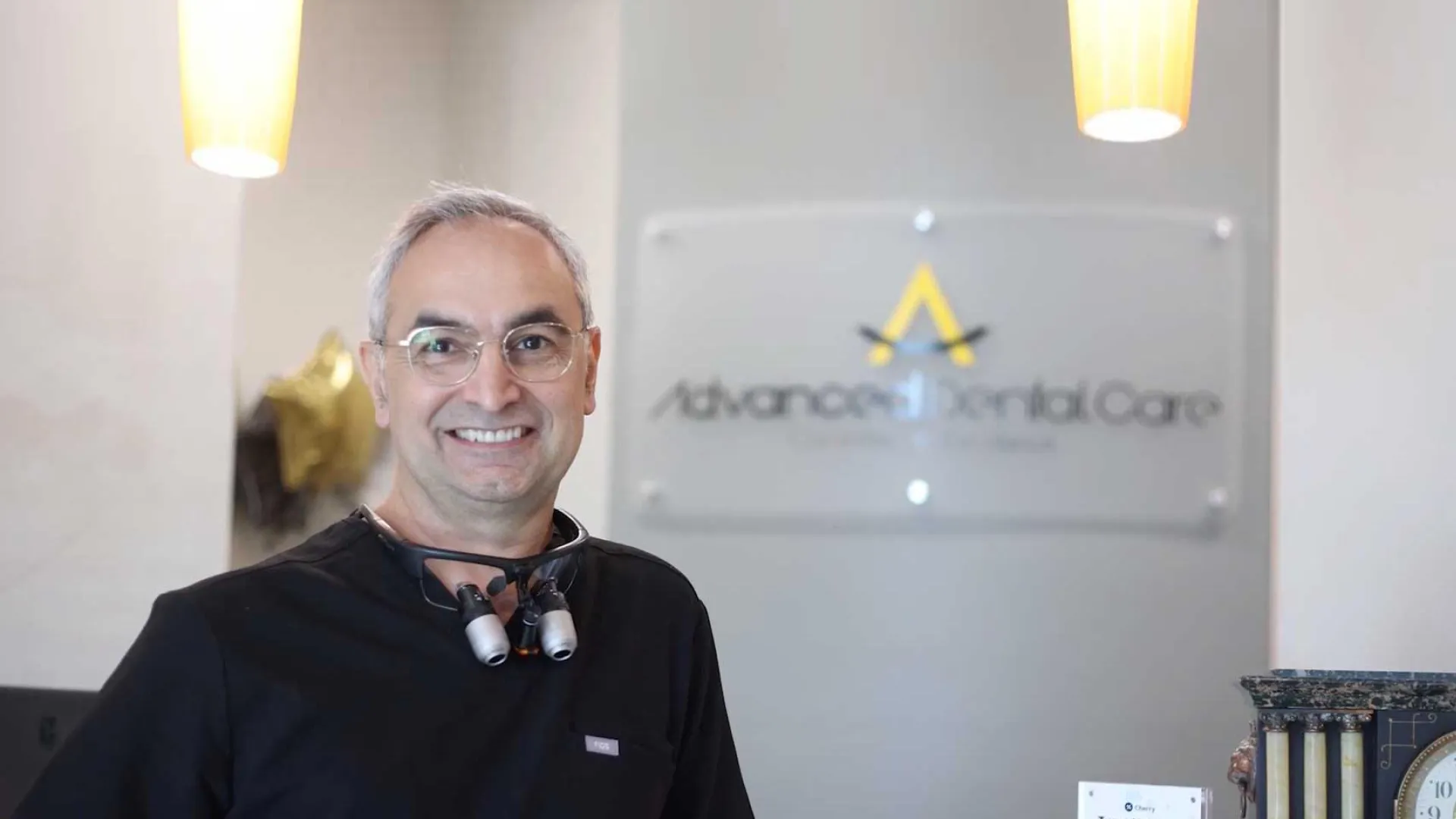 Smiling male dentist wearing magnifying loupes standing in a modern dental clinic reception area.