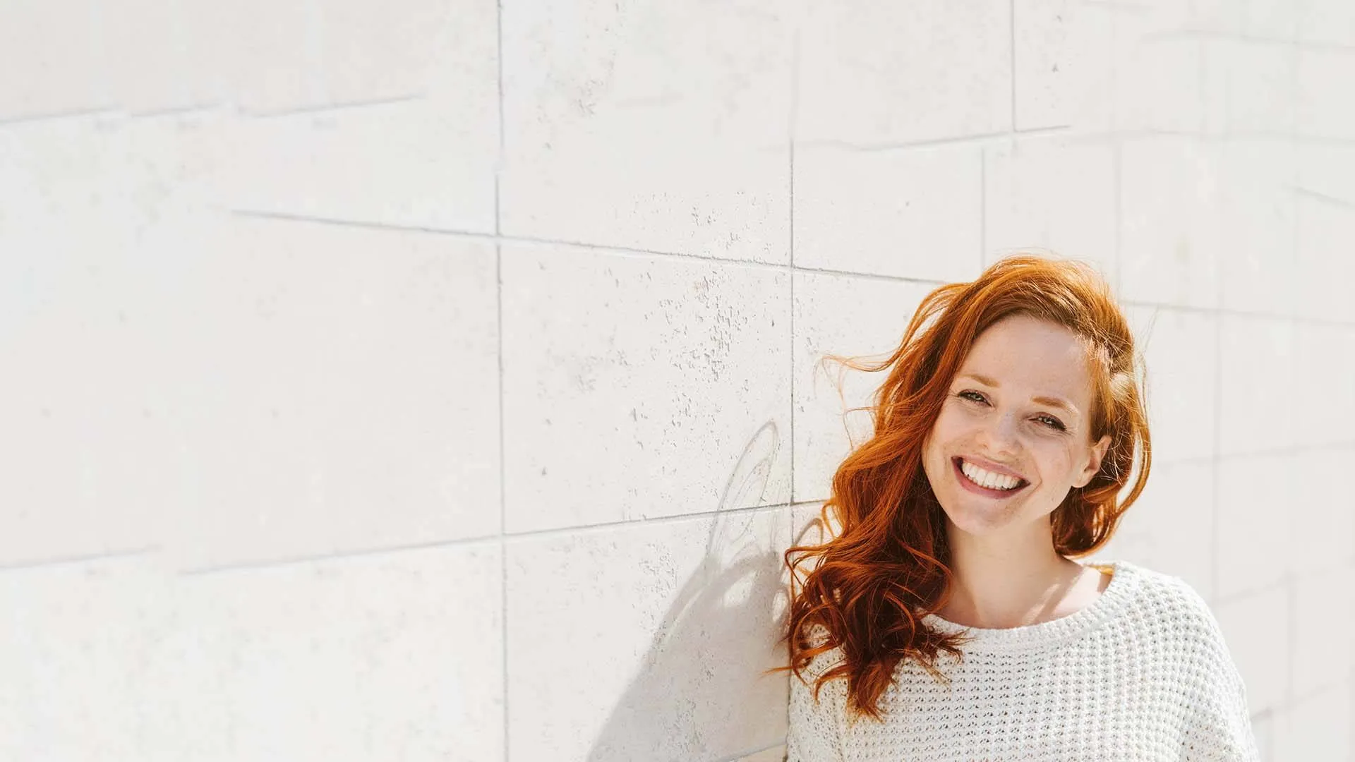 Smiling red-haired woman wearing a white sweater leaning against a light stone wall outdoors