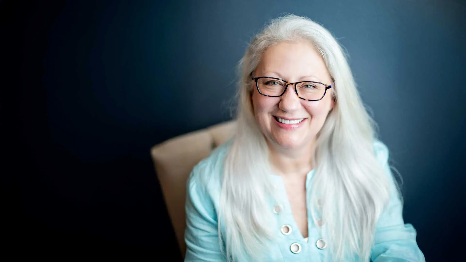 Smiling mature woman with long white hair and glasses wearing a light blue blouse against a dark background