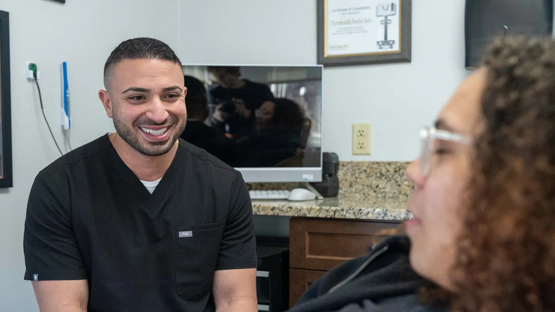 Smiling male healthcare professional in black scrubs talking to female patient in a medical office.