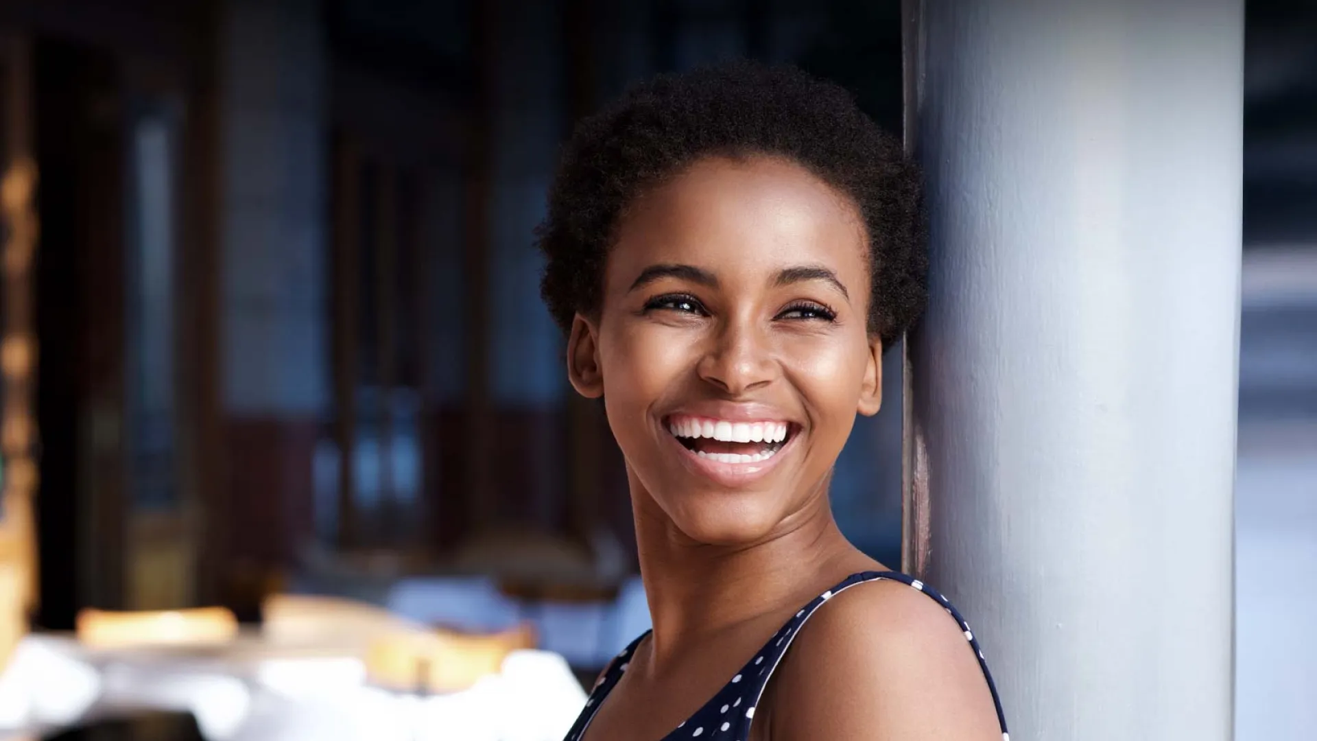 Smiling woman in polka dot dress, leaning against a pole in a cozy outdoor setting.