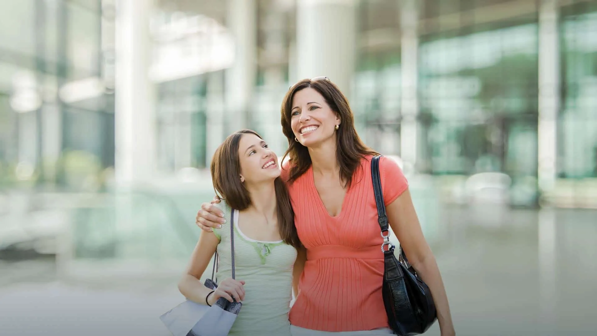 Smiling mother and daughter walking arm in arm outside a modern glass building on a sunny day