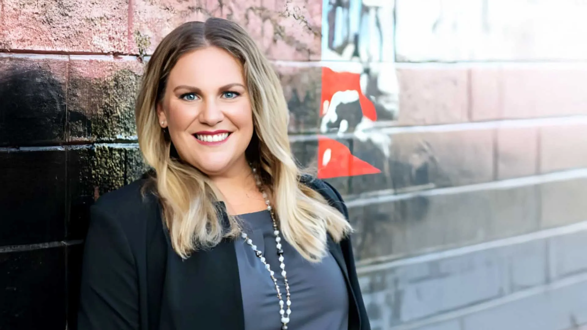 Smiling woman with blonde hair wearing black blazer and pearl necklace posing against a painted brick wall with red and black art.