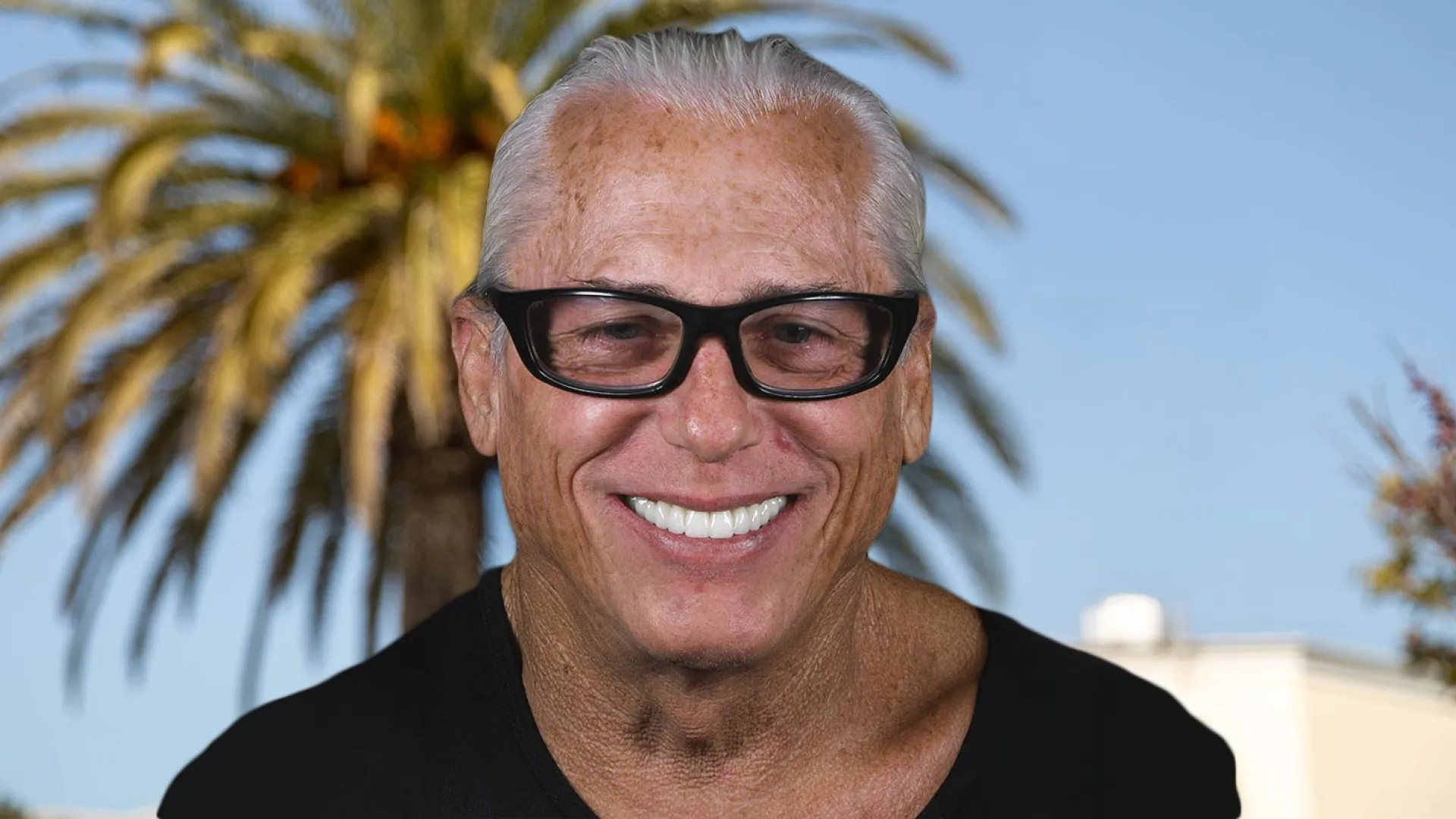 Smiling man with glasses against a palm tree background, showcasing a sunny atmosphere.