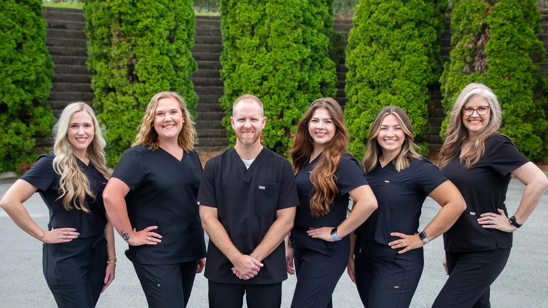 Group of six healthcare professionals in black scrubs standing outdoors in front of green bushes smiling at camera.