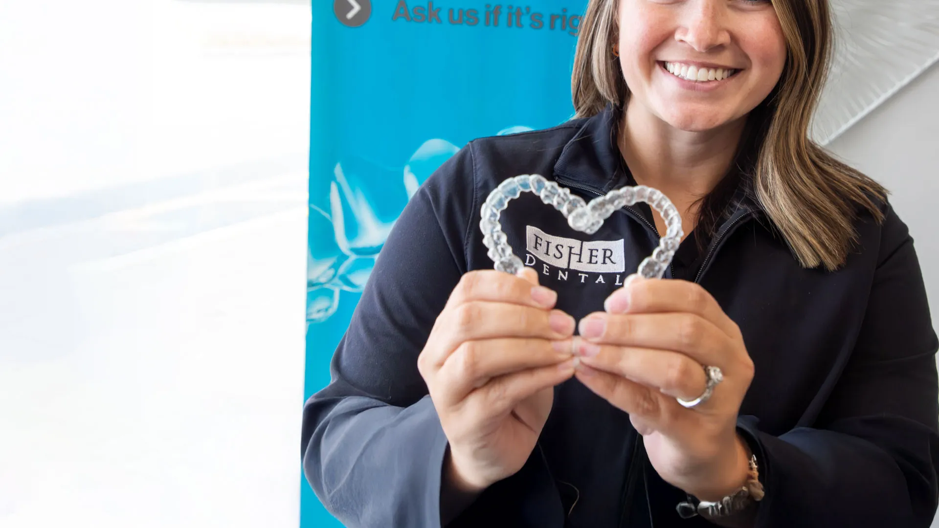 Woman smiling and holding clear dental aligners shaped as a heart with Fisher Dental logo in background