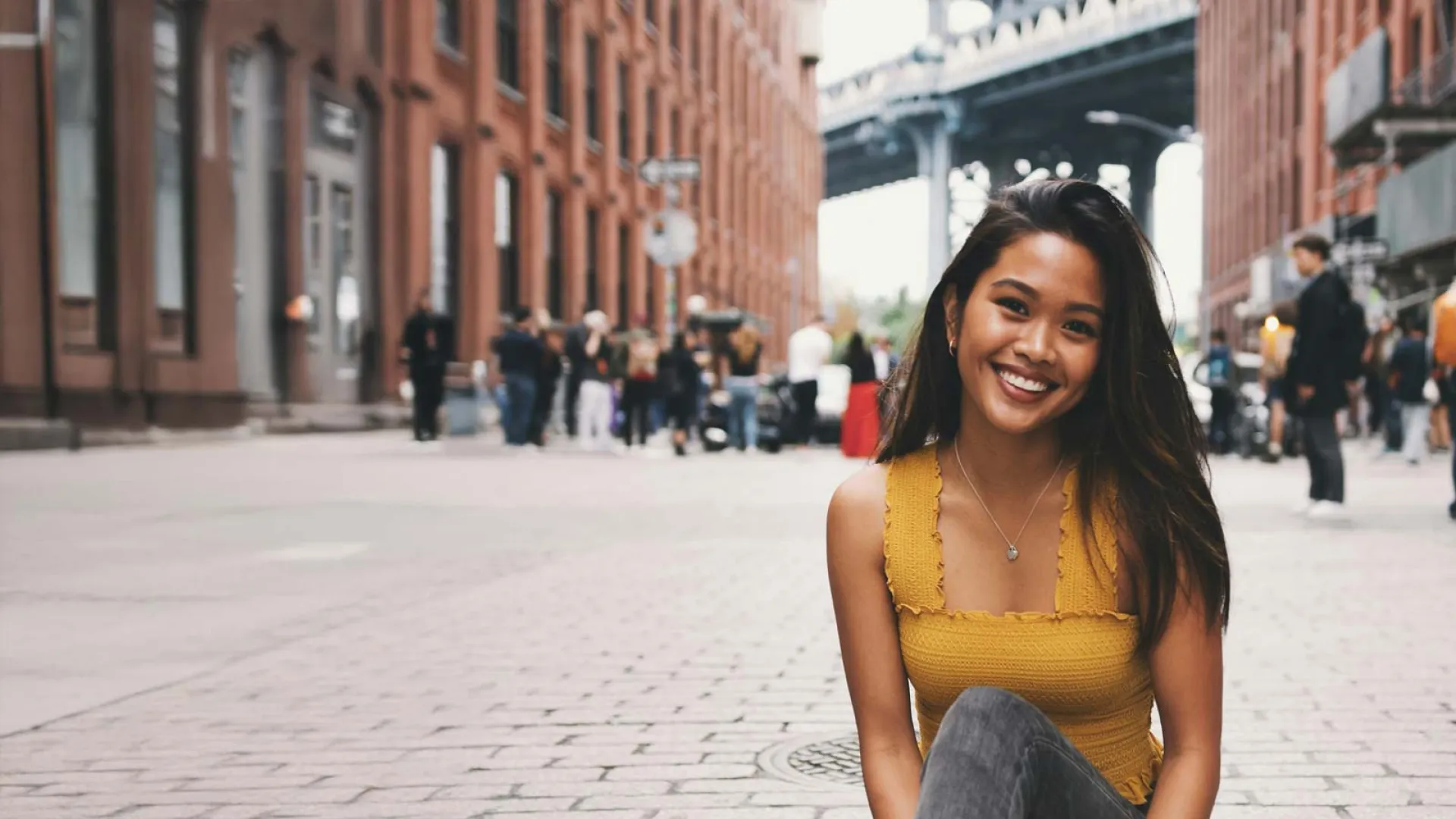 Smiling young woman in yellow top sitting on urban street with brick buildings and bridge in background.