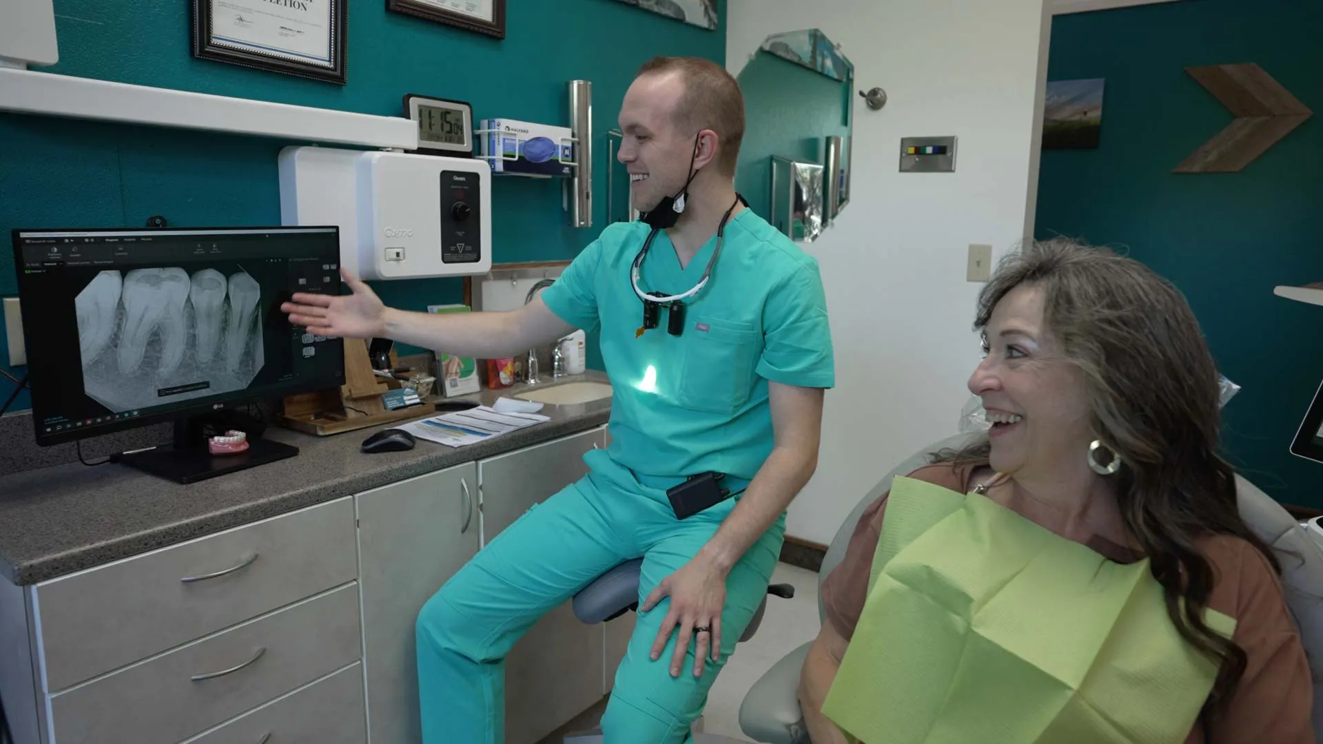 Dentist in teal scrubs showing a dental X-ray on monitor to a smiling female patient in a dental office.