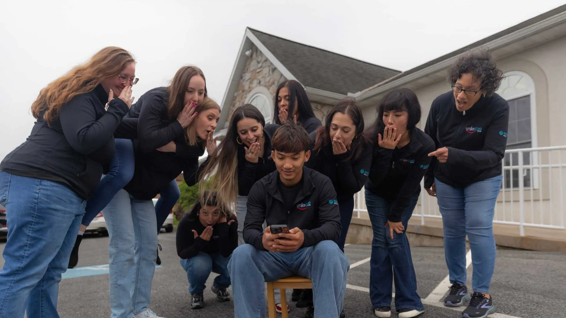 Group of people excitedly looking at a phone held by a seated man outdoors near a building with stone facade.