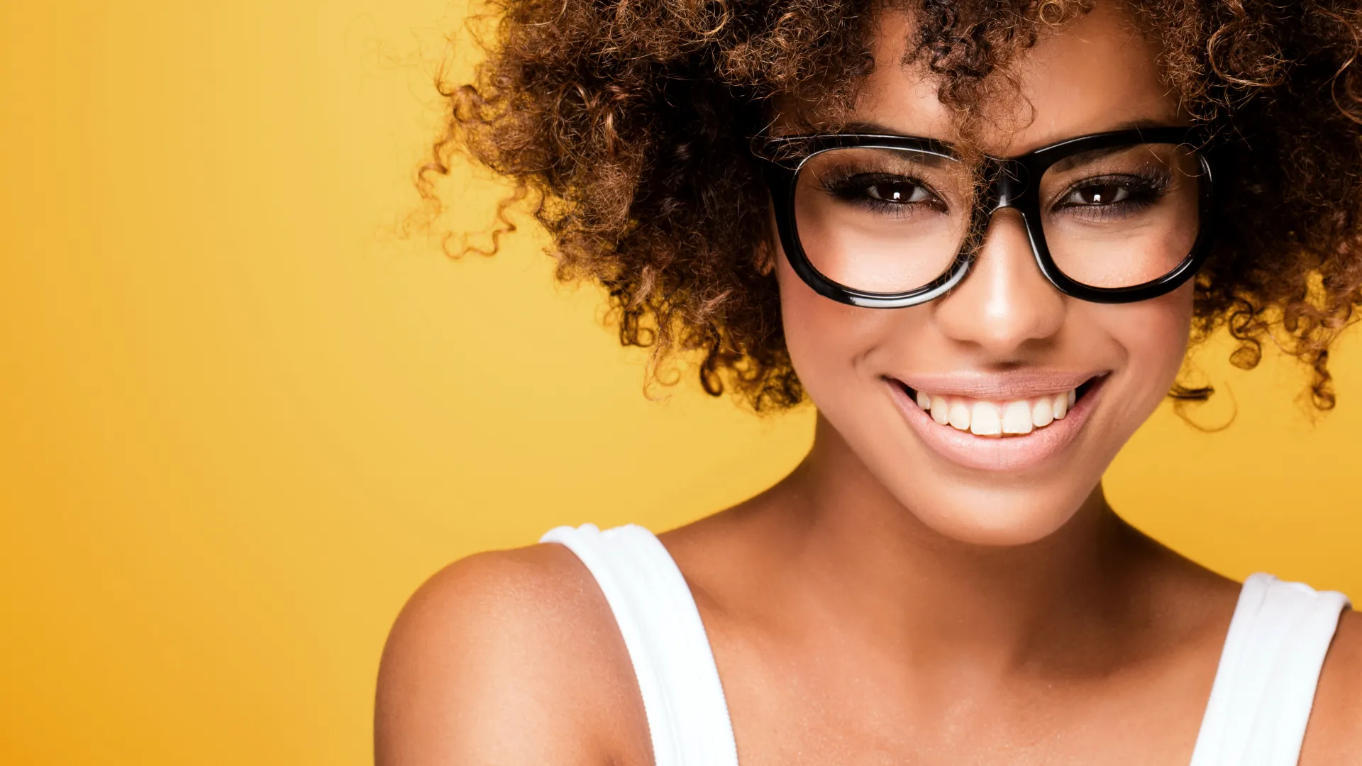 Smiling woman with curly hair and black glasses against a yellow background wearing a white top
