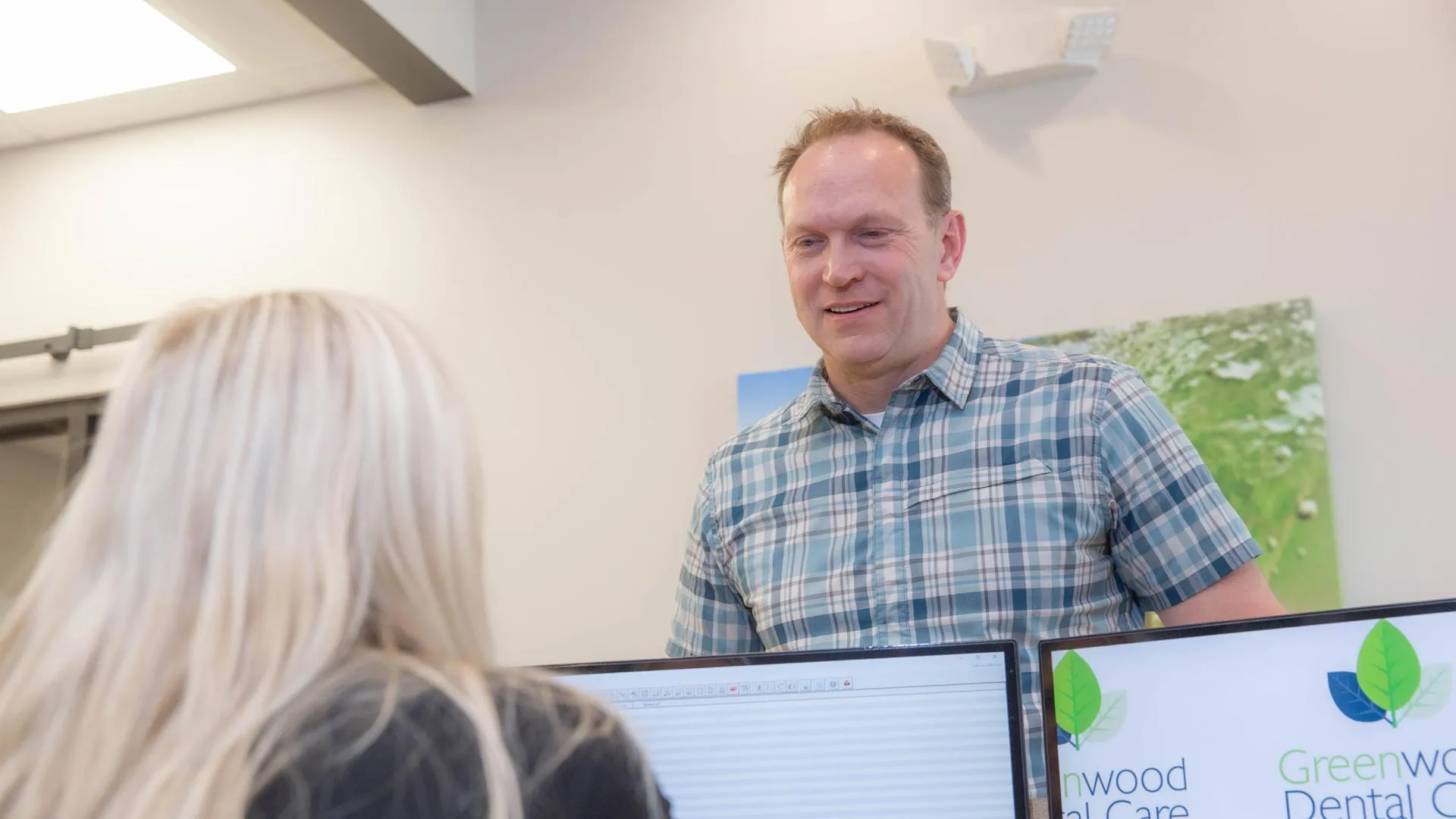 Man in plaid shirt speaking with woman at dental office reception with Greenwood Dental Care logo visible