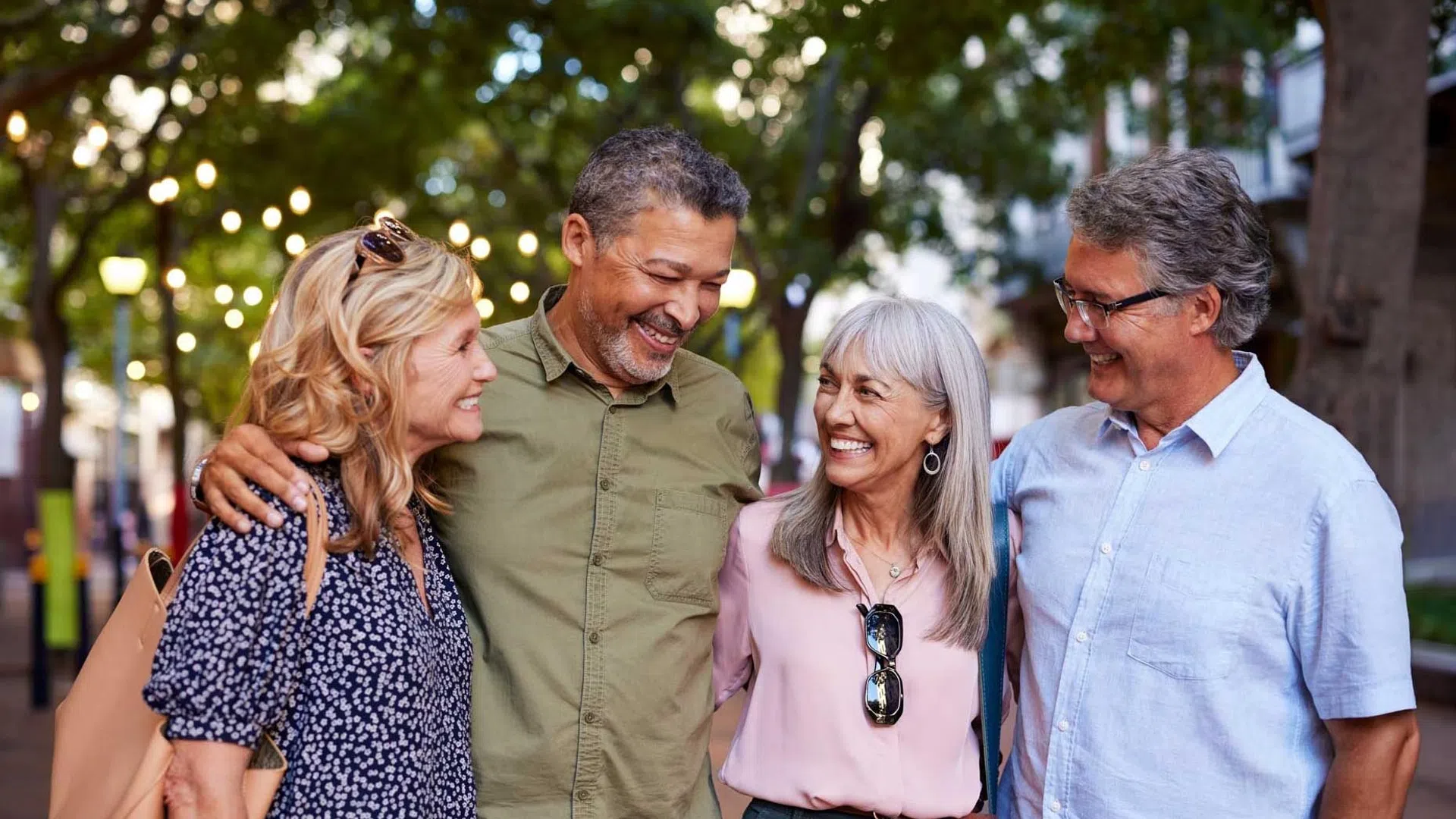 Four middle-aged friends smiling and walking together outdoors under string lights and green trees in the evening.