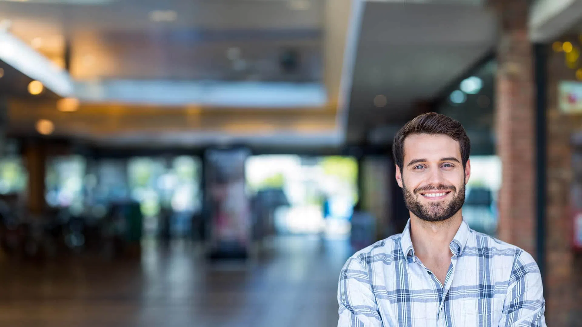 Smiling bearded man in plaid shirt standing with arms crossed in a bright modern indoor space.