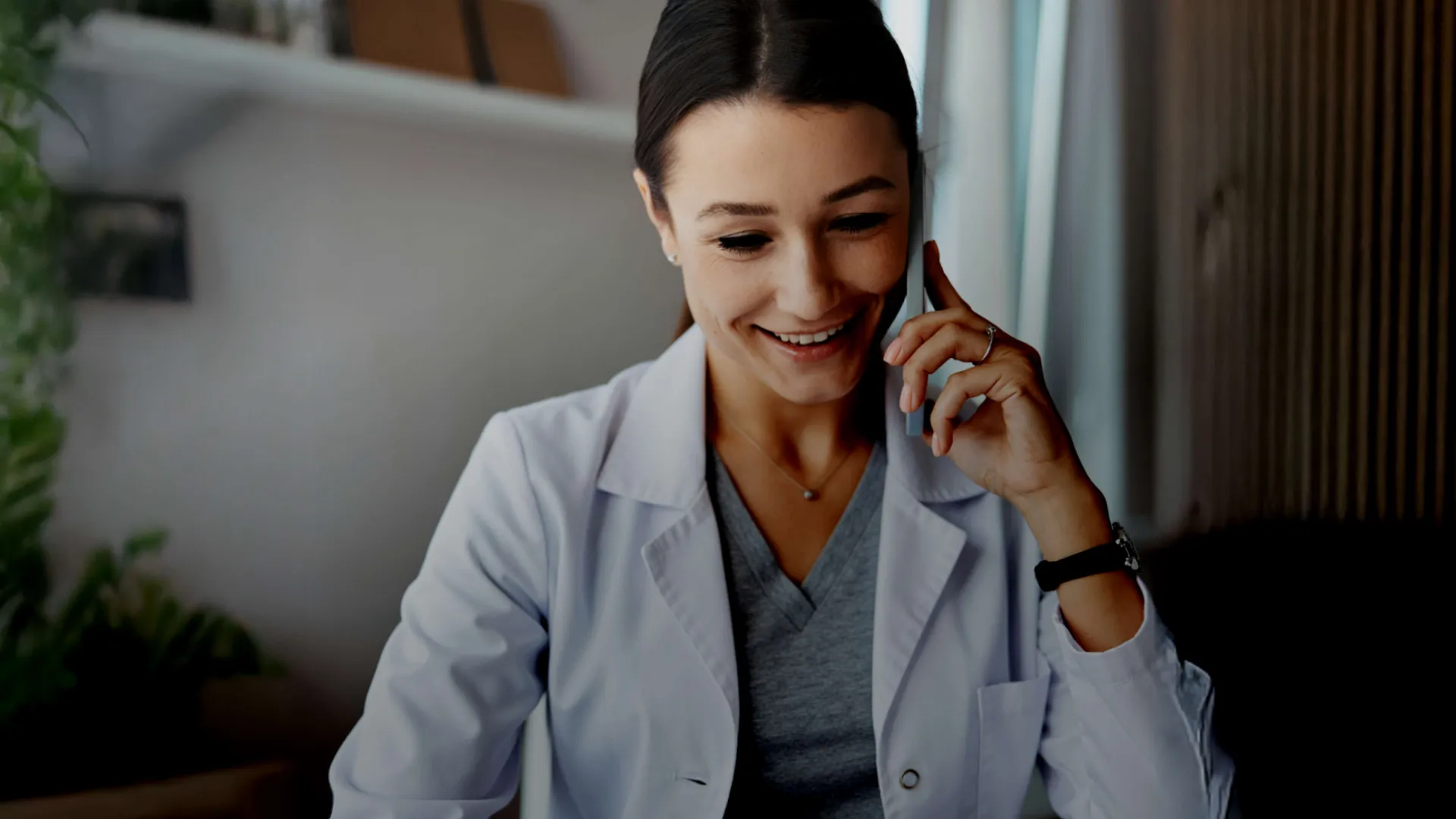 Smiling female doctor in white coat talking on phone in a cozy office setting with natural light.