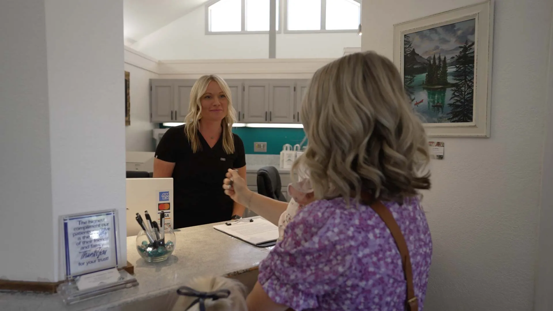 Receptionist assisting a woman with a baby at a clinic front desk inside a bright reception area.