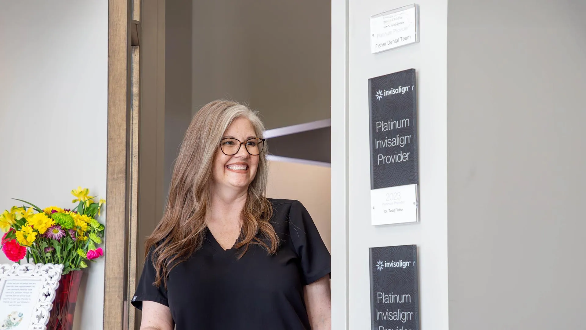 Smiling female dental professional standing in doorway near Platinum Invisalign Provider signs and floral arrangement.