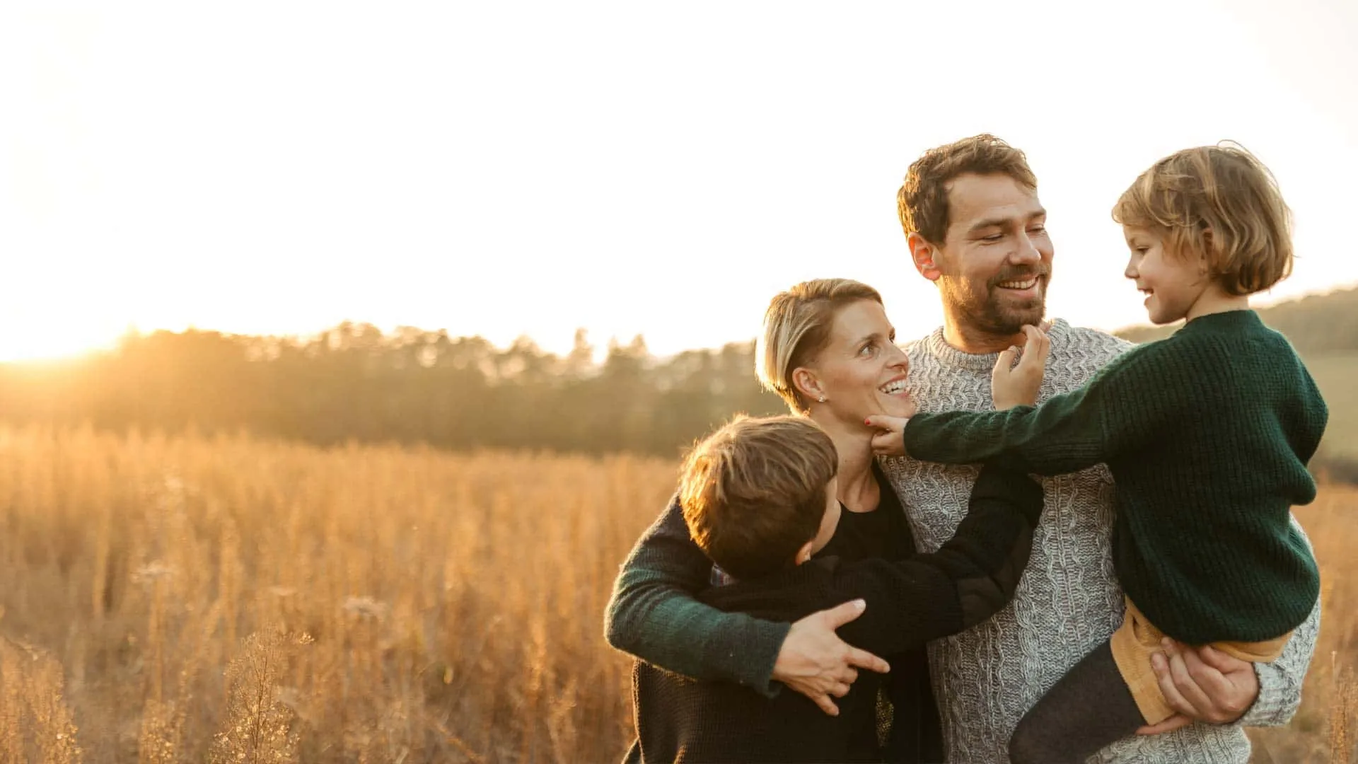 Happy family of four embracing each other in a sunlit field at sunset.