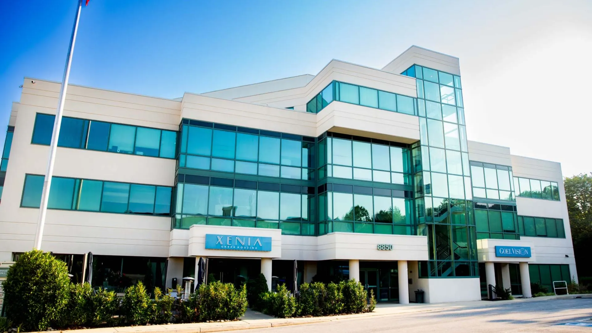 Modern three-story office building with large glass windows under clear blue sky and surrounding greenery