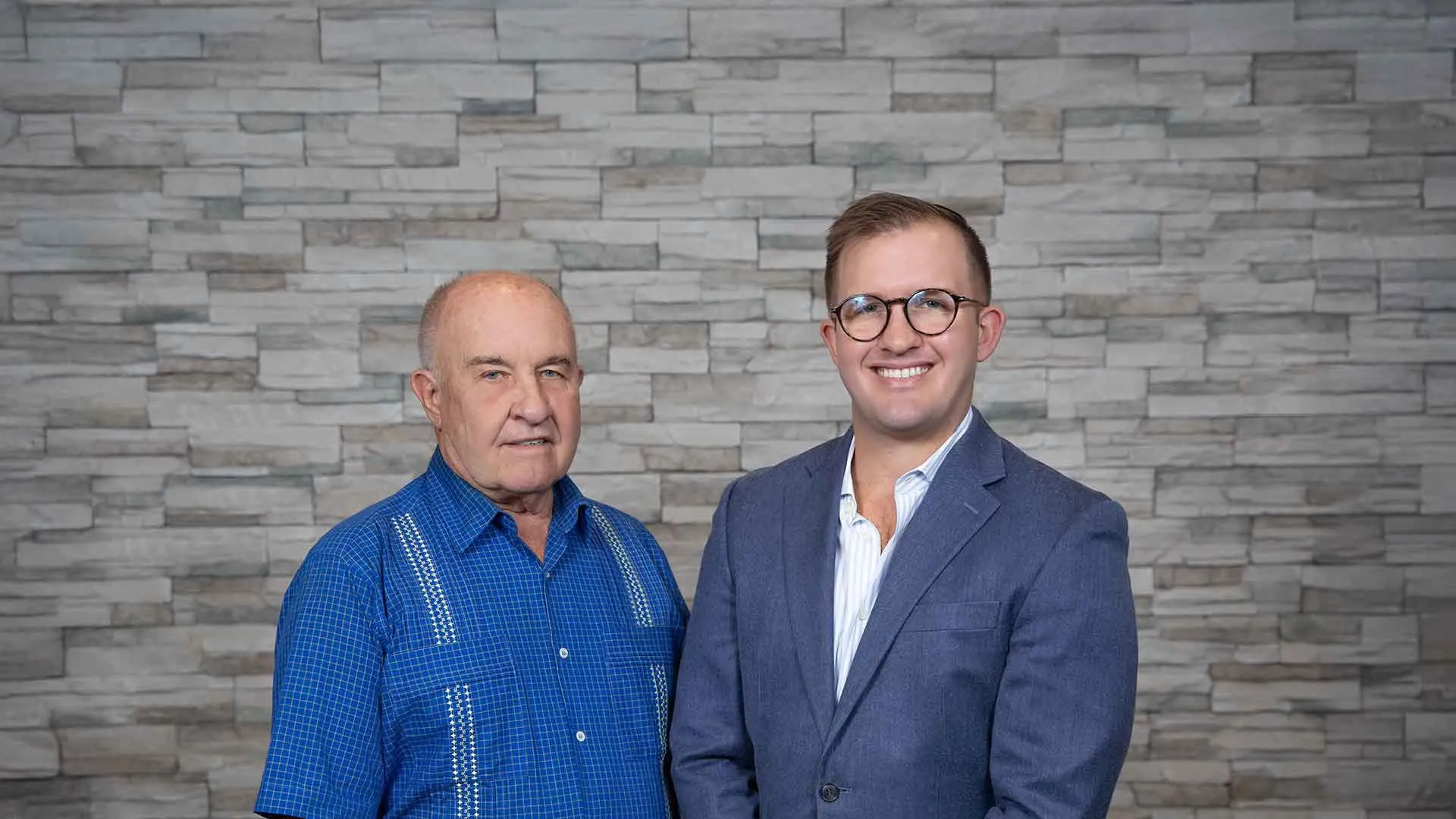 Older man in blue shirt and younger man in suit smiling, standing against gray stone wall background.