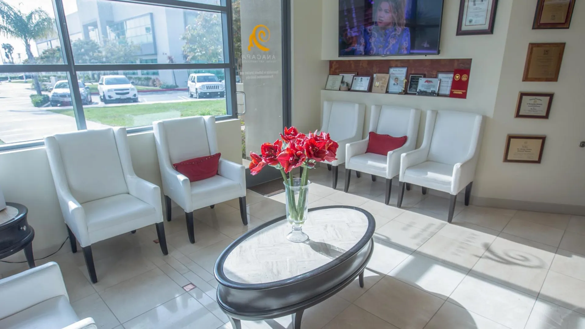 Modern office reception area with white chairs, a vase of red flowers, and large windows.