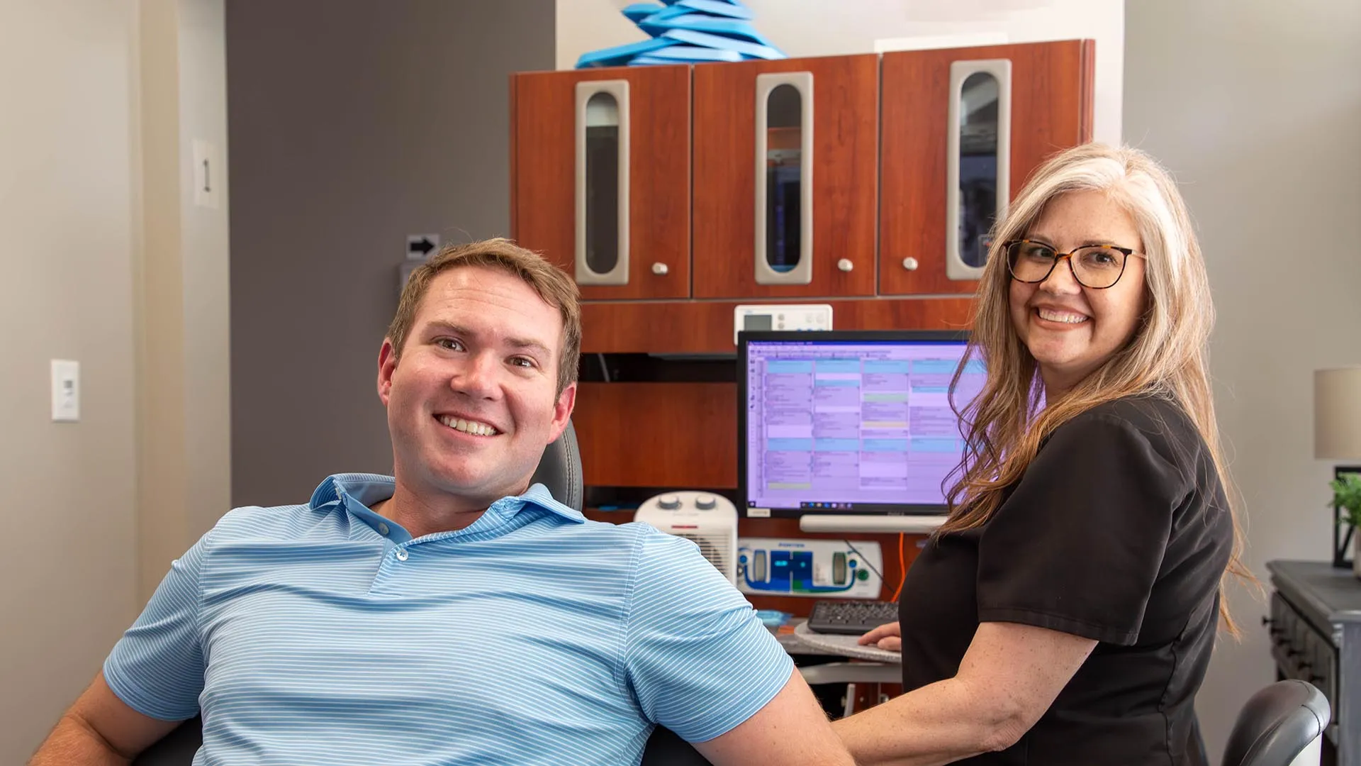 Smiling man in blue polo sitting in dental chair with female dental hygienist in black scrubs in bright office