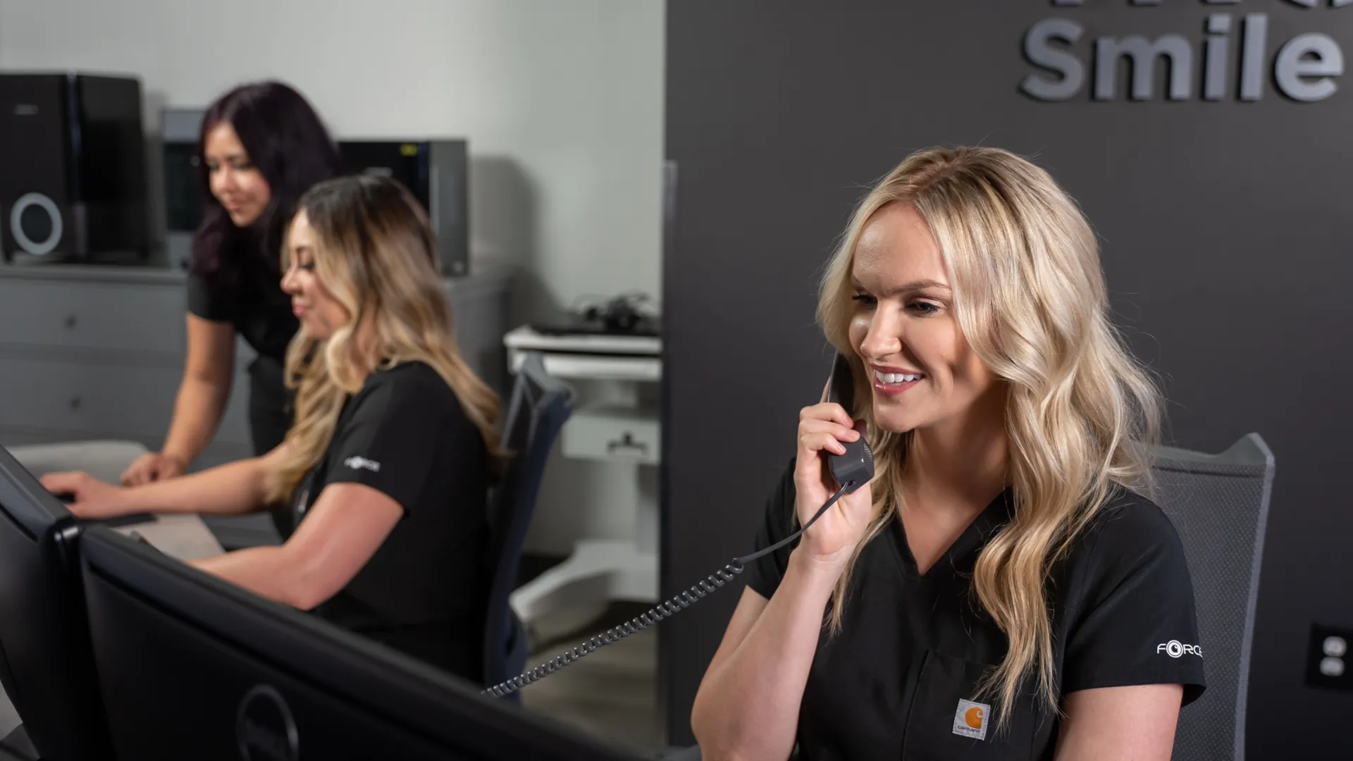 Three women working at computer desks in a modern office, one smiling while talking on the phone.
