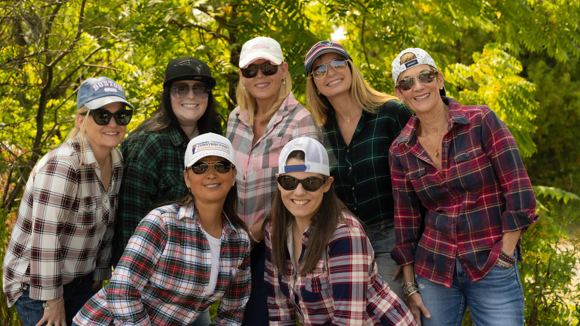 Group of seven women wearing sunglasses and plaid shirts smiling outdoors with green trees in background