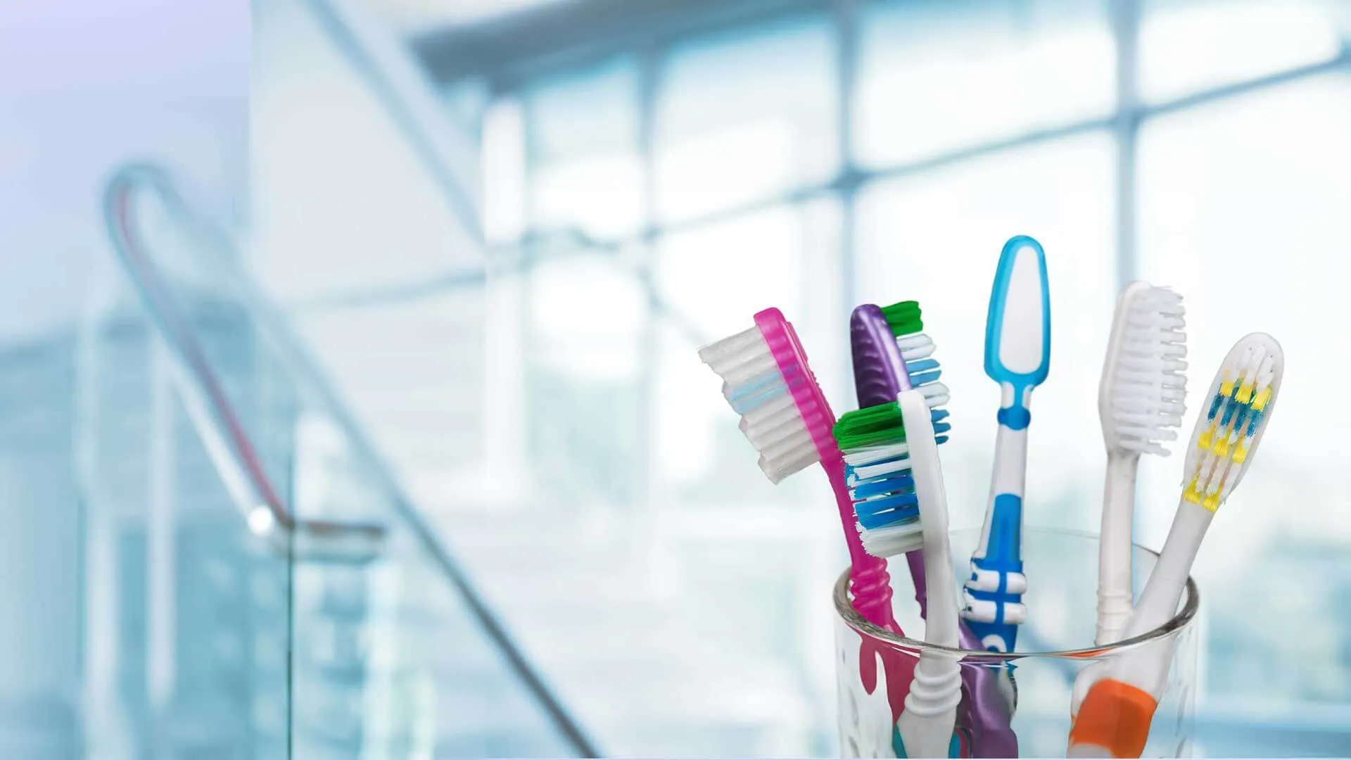 Assorted colorful toothbrushes standing upright in a transparent glass with blurred indoor background.