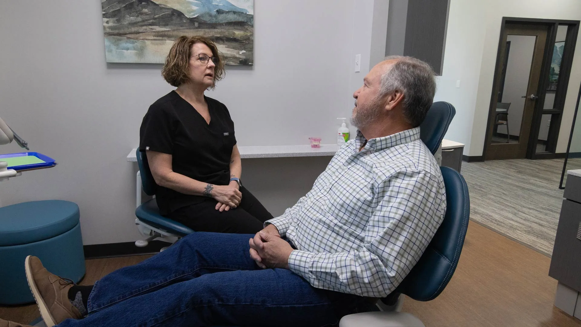 Dentist in teal scrubs showing a dental X-ray on monitor to a smiling female patient in a dental office.