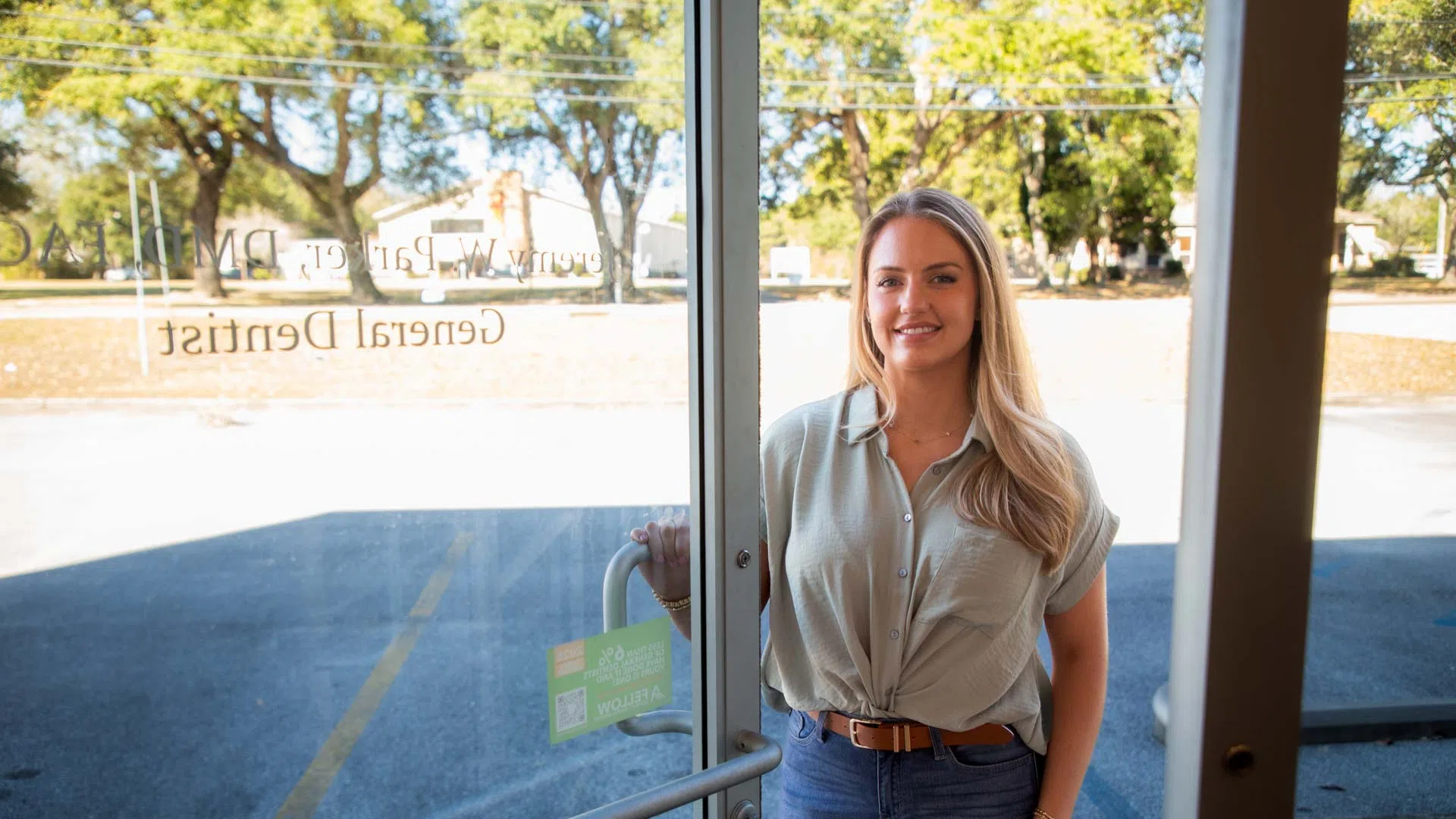 Smiling woman holding door open standing outside a dental office with trees and parking lot in background