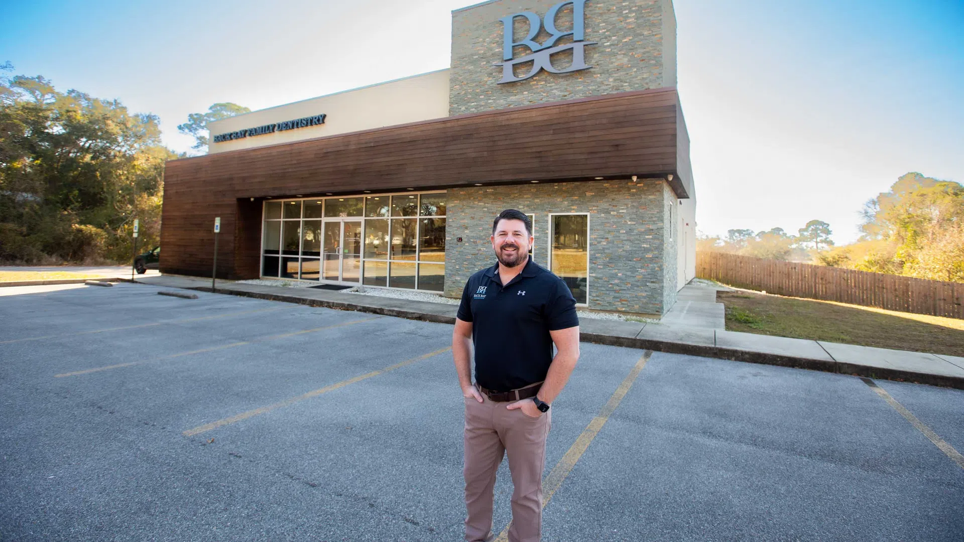 Man standing in front of a modern dentist office building with stone and wood exterior under clear sky.