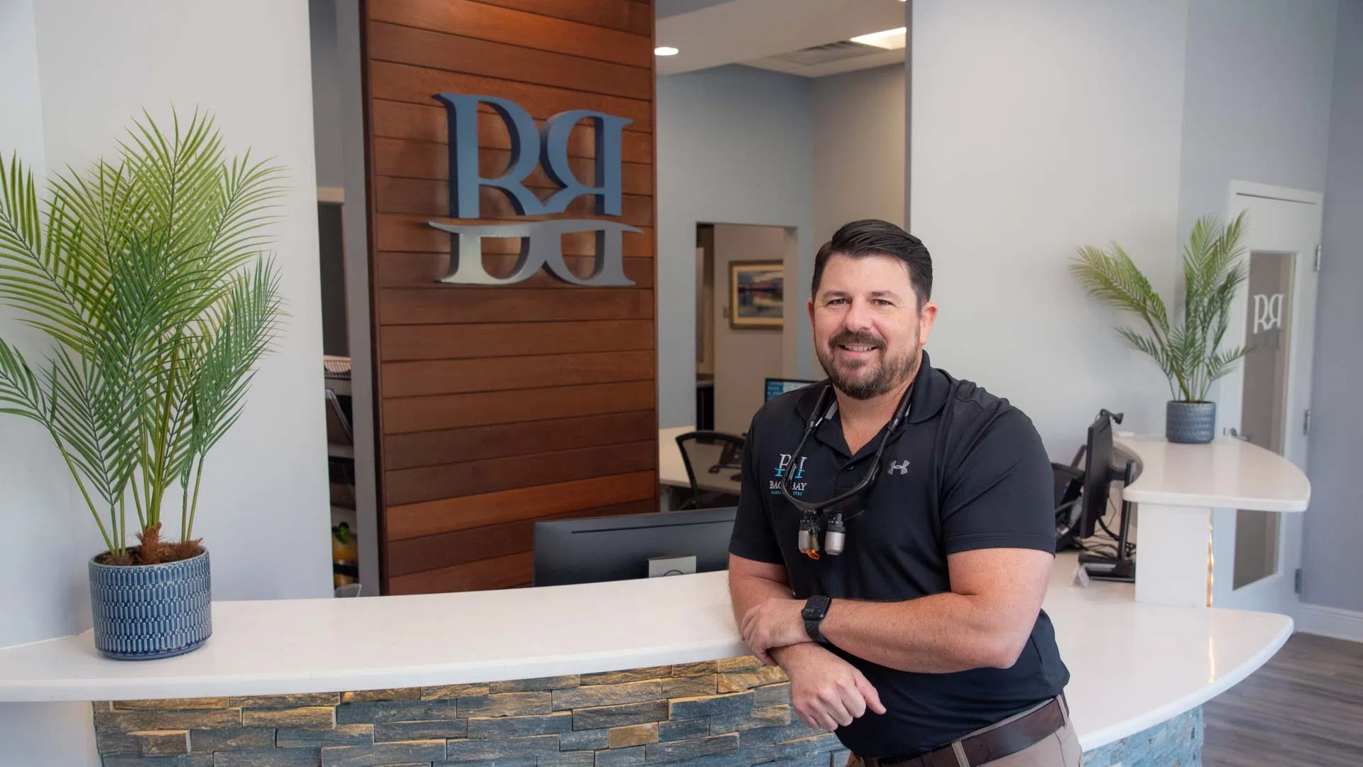 Smiling dentist leaning on modern reception desk with stone facade and plants in a dental office lobby.