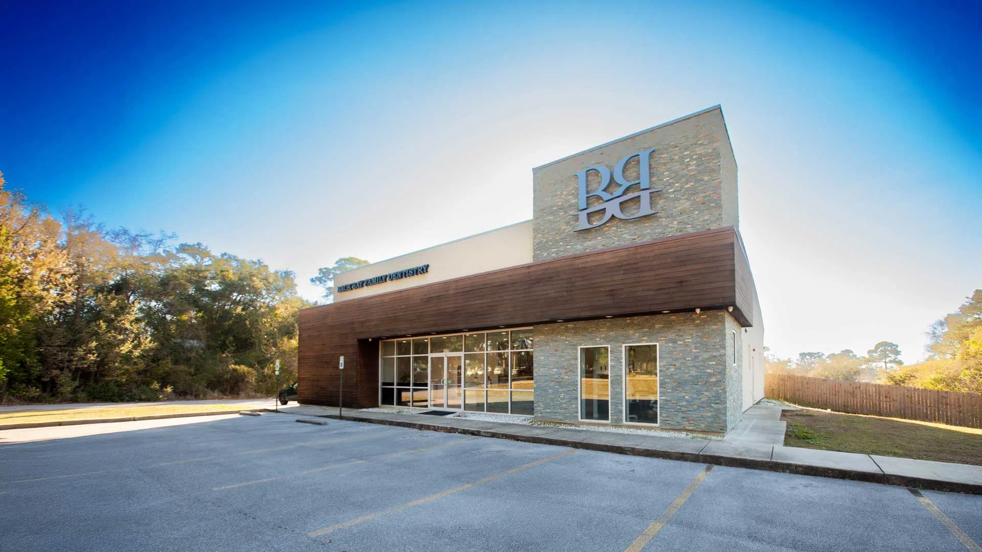 Modern building with stone and wood exterior featuring large windows and a parking lot under a clear blue sky