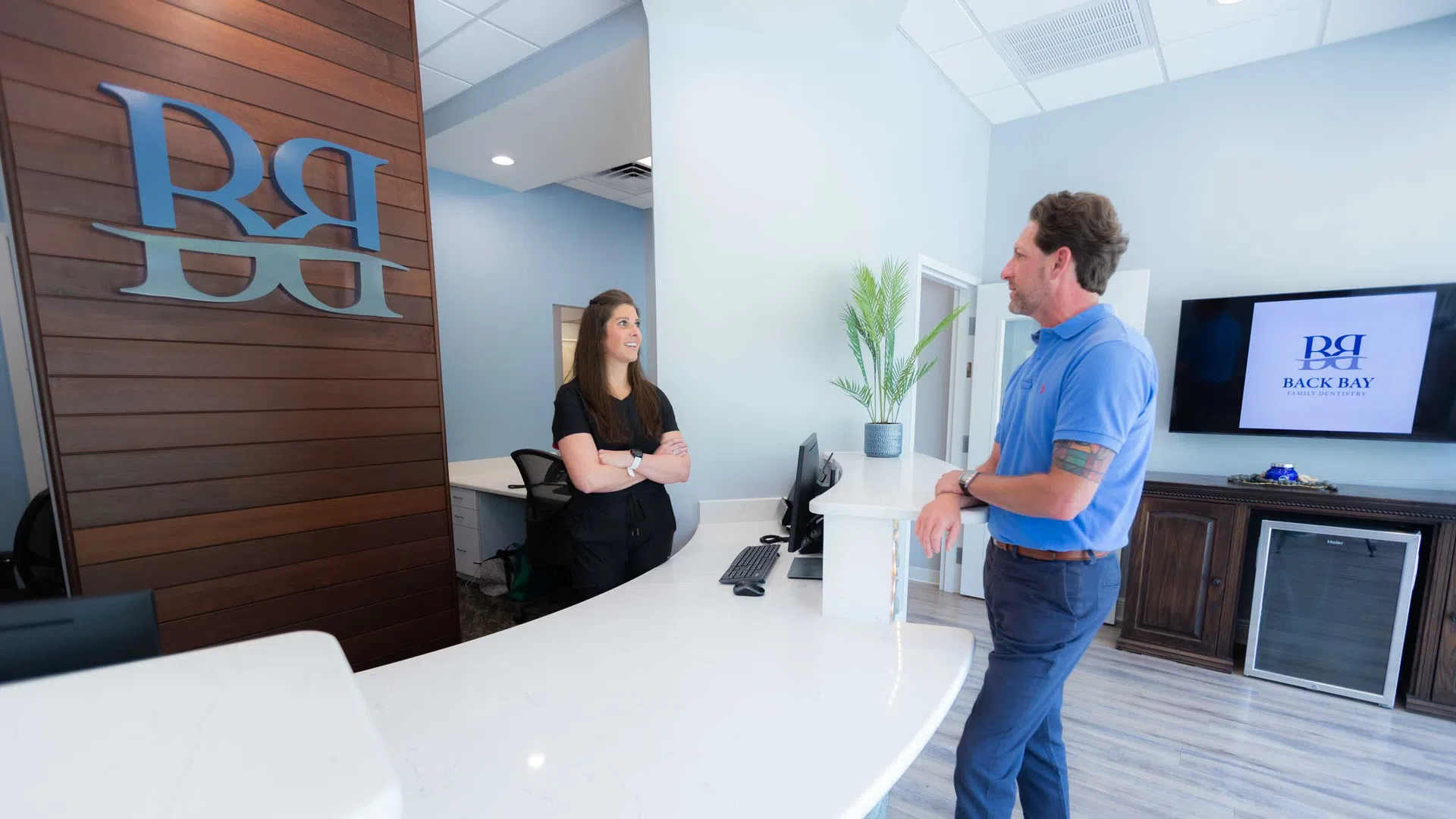 Reception area with two people conversing, modern white desk, wood panel wall with logo, and flat screen displaying logo.