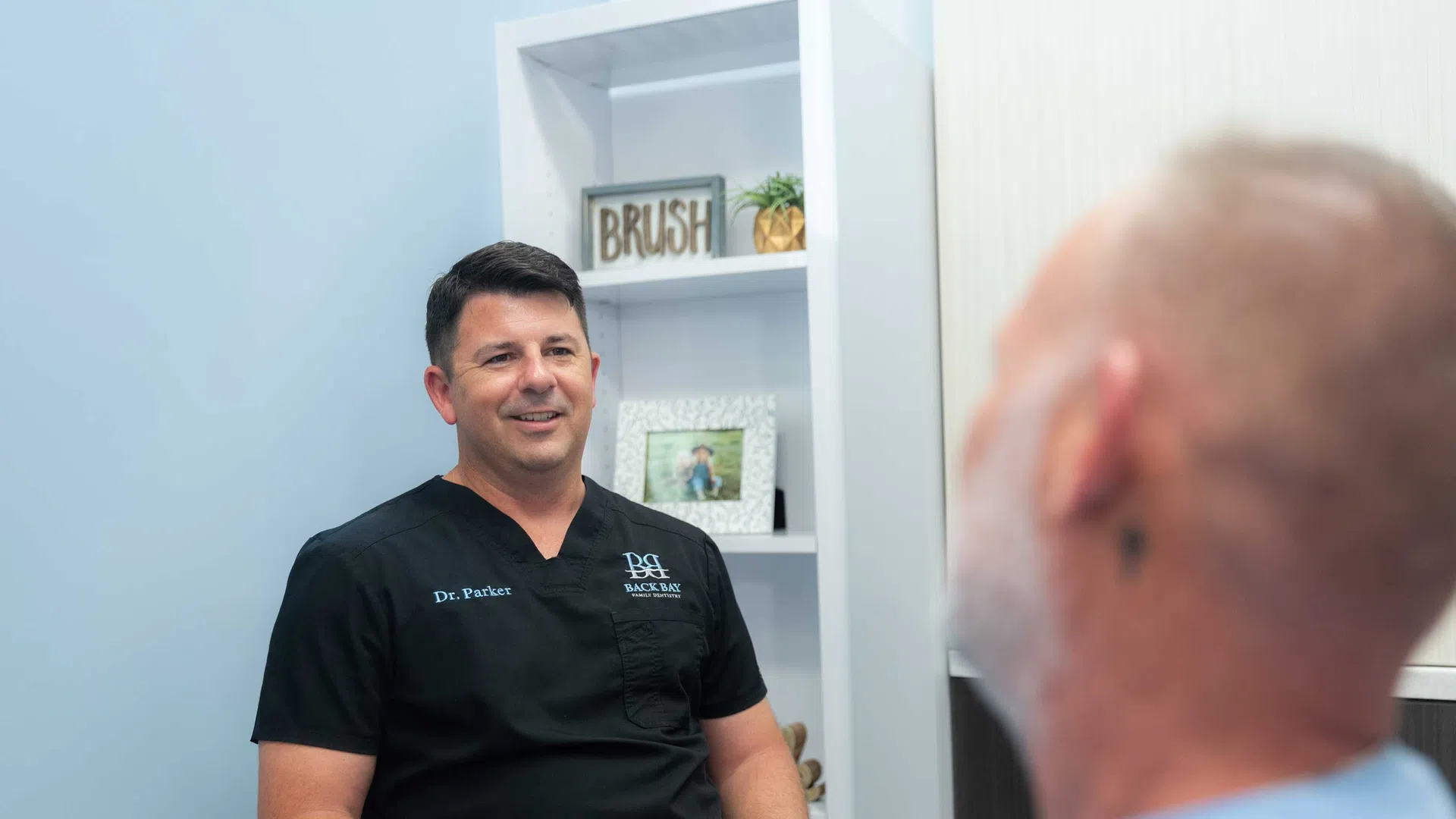 Doctor in black scrubs consulting with a patient in a modern medical office with white shelves.