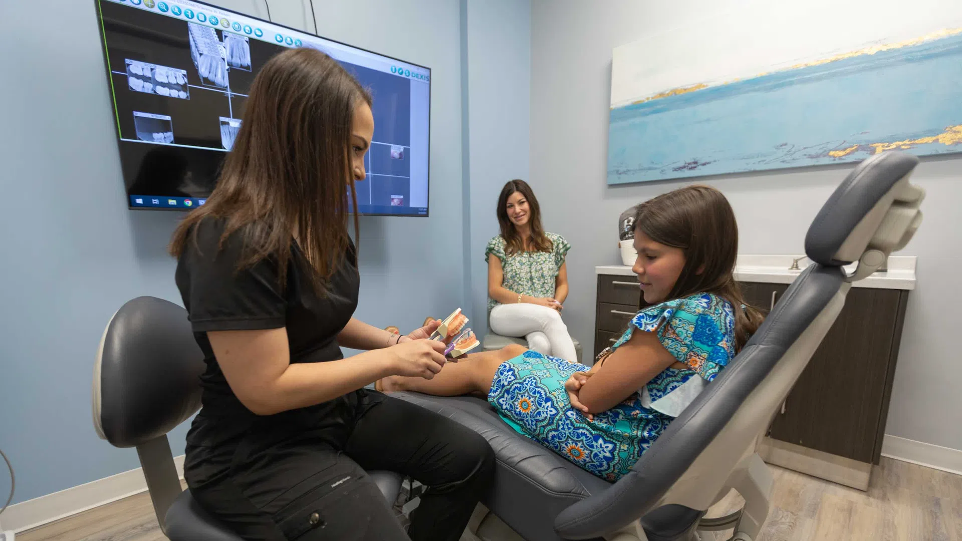 Dentist showing a young girl a dental model in a modern clinic with mother watching nearby.