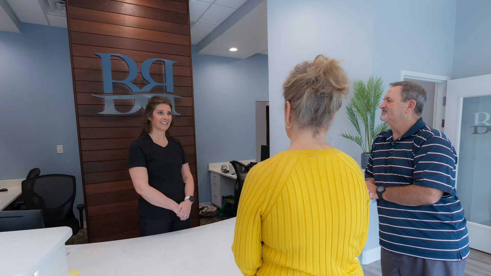 Receptionist talking to a woman in a yellow sweater and a man in a striped shirt at a modern front desk.