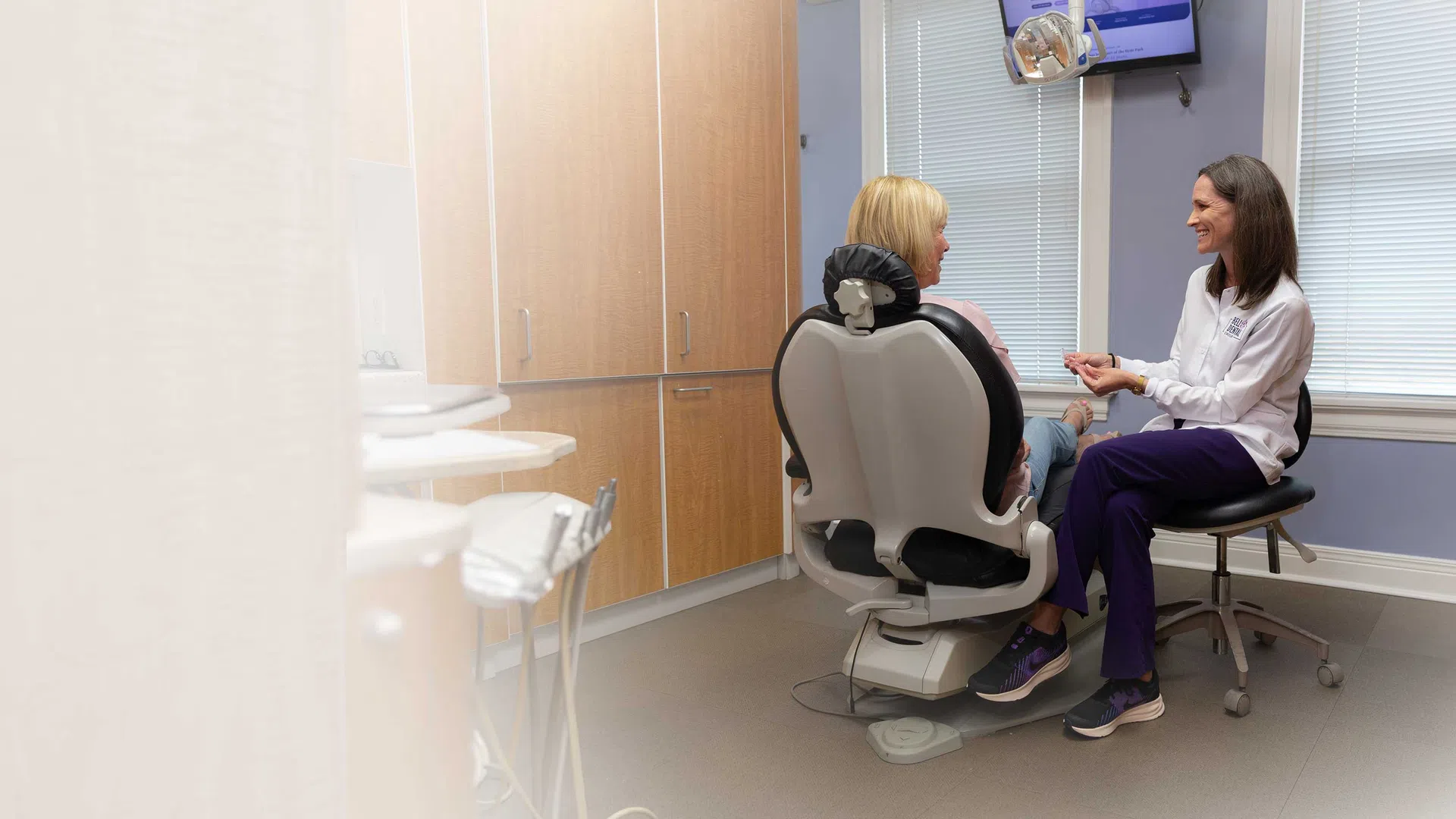 Dentist consulting with patient in dental office, both smiling and seated by window with blinds.